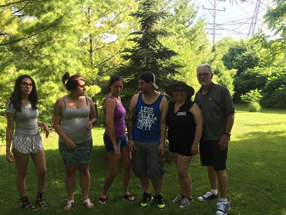 Six people stand in a row on a sunny grassy lawn in a park wearing casual summer clothes, posing for a photo.