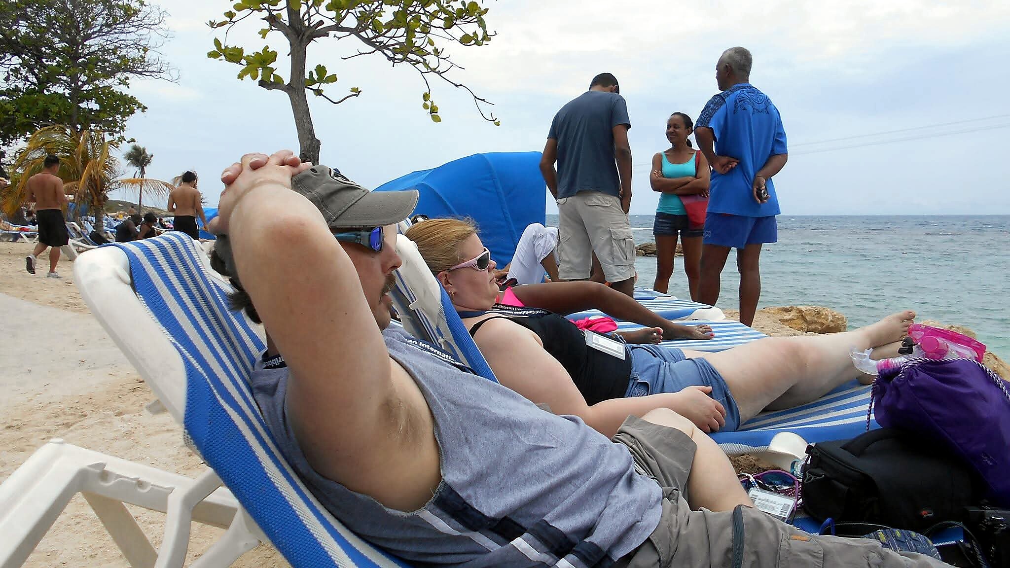 Sunbathers on blue-striped lounge chairs at a sandy beach, with people standing and chatting near the water in the background.