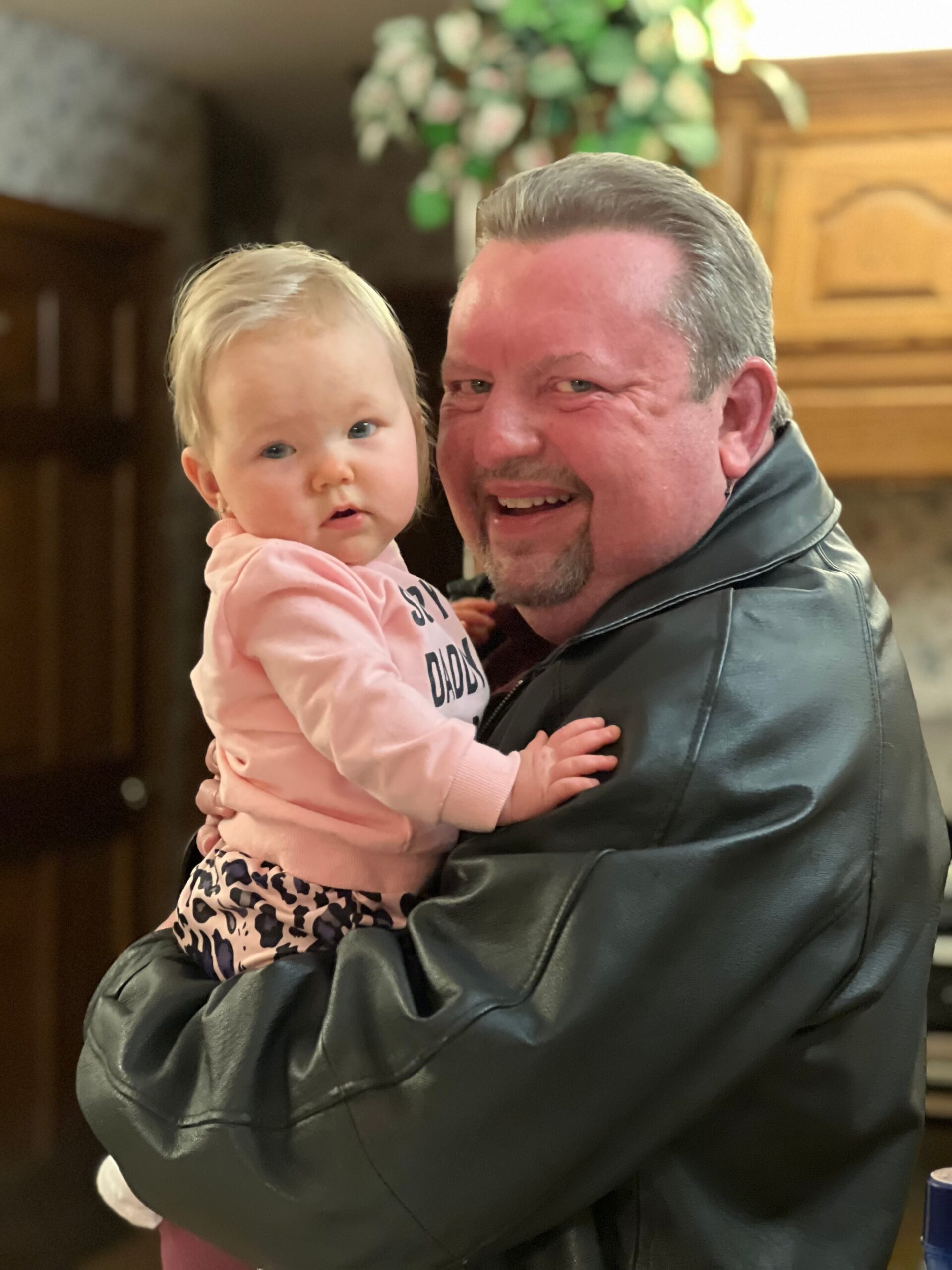 A man in a dark jacket smiles while holding a young child in a pink outfit, both looking at the camera indoors.