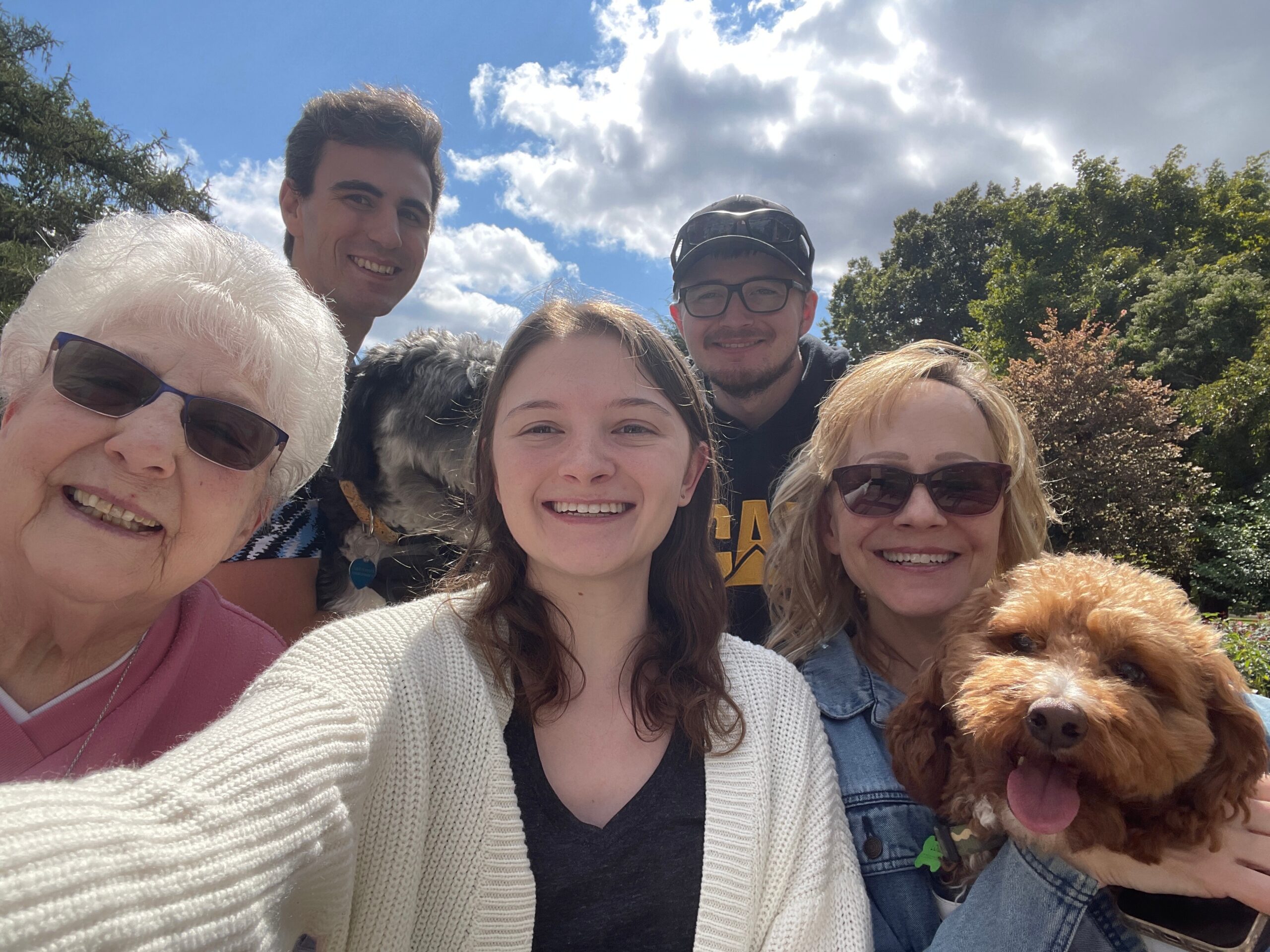 Group selfie outdoors with smiling people and a brown dog in the foreground under a blue sky and trees outside.