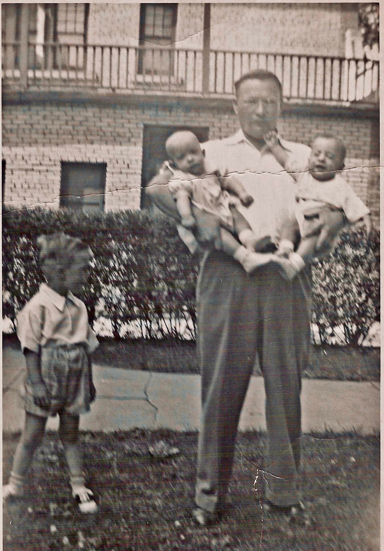 A man stands outside holding two babies, one in each arm, with a young boy nearby by shrubs; brick building and a fence behind them.