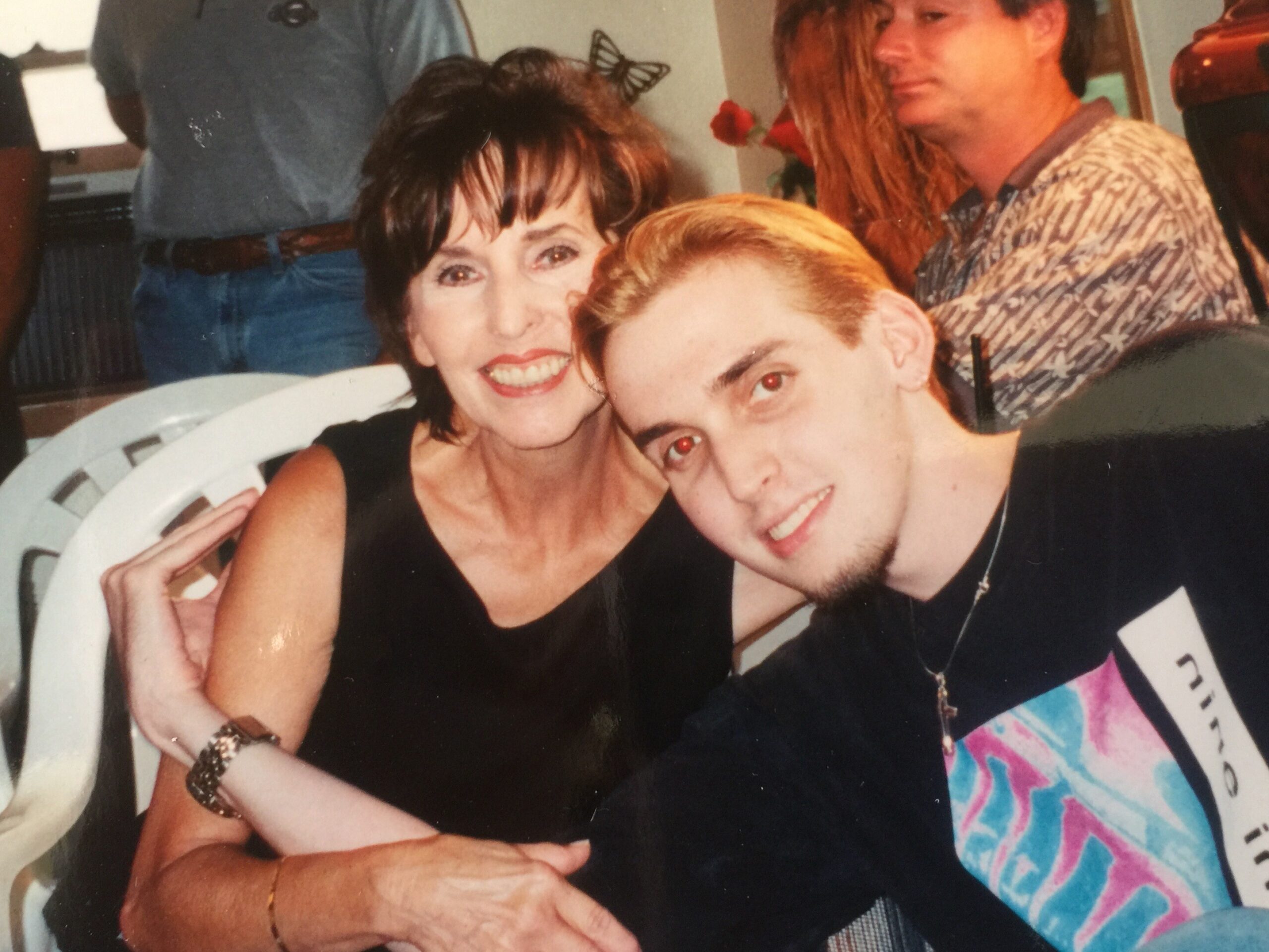 Two friends posing closely for a photo: a smiling woman with short dark hair in a black top and a young man with blond hair leaning in, both looking at the camera.