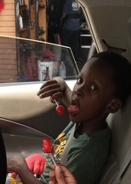 Young child in a car seat inside a car, holding red lollipops and licking one.