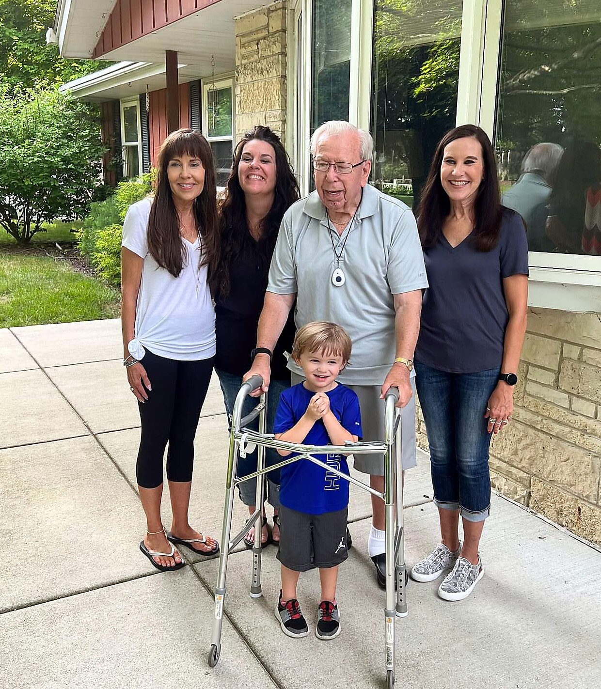 A smiling elder man with a walker stands beside a young boy with a walker, flanked by four adults outside a house.
