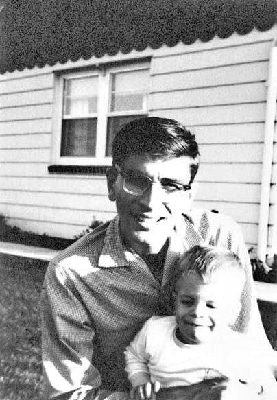 Man with glasses holding a smiling toddler outdoors in front of a suburban house (black-and-white photo).