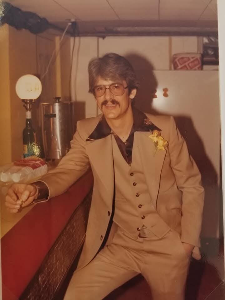 Man with gray hair and mustache in a beige 3-piece suit and yellow boutonniere leaning at a bar, holding a drink. Variants: retro vibe in a dimly lit bar setting.