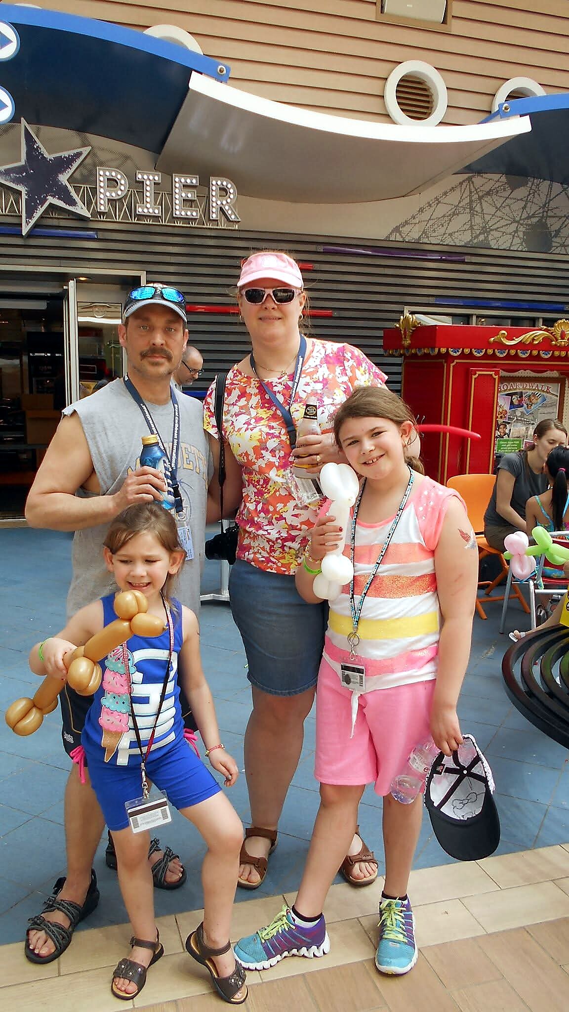 Family of four posing for a photo at a daytime event, each holding balloon animals and wearing lanyards in a colorful outdoor venue.