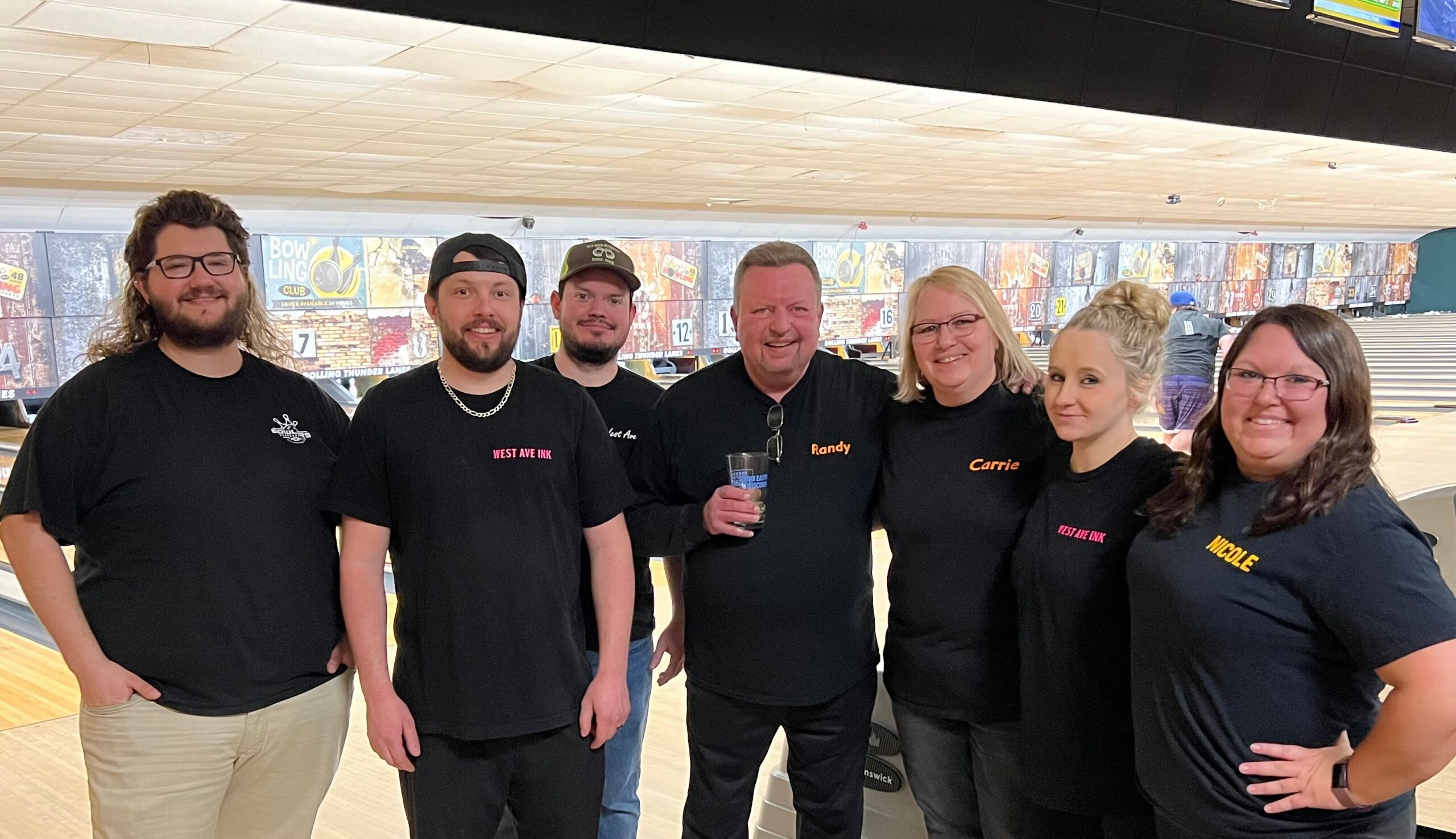 Seven people in black shirts pose together at a bowling alley, lanes and scoreboards in the background.