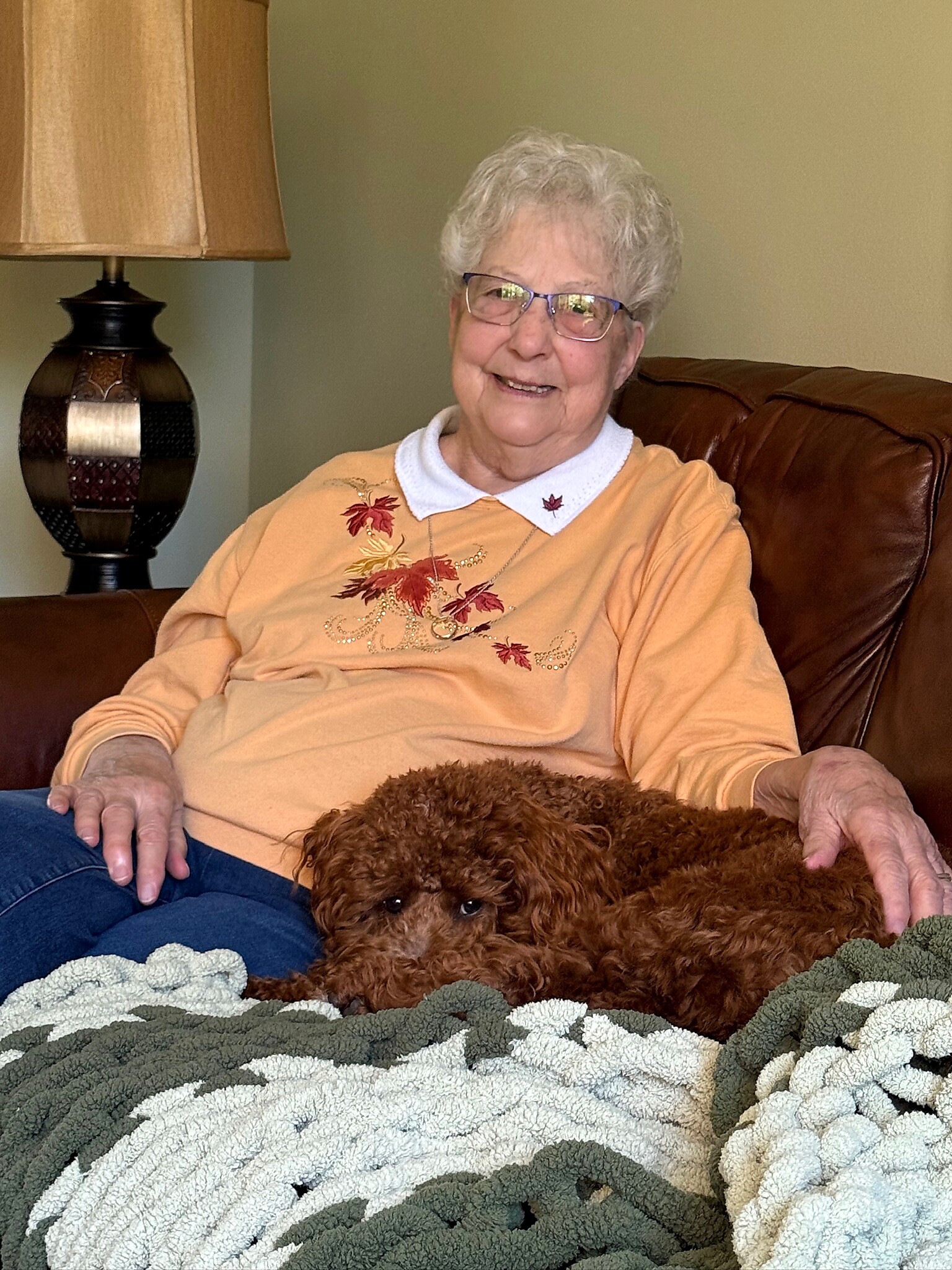 Smiling elderly woman with glasses sits on a brown leather couch, with a fluffy brown dog resting in front of her on a blanket.