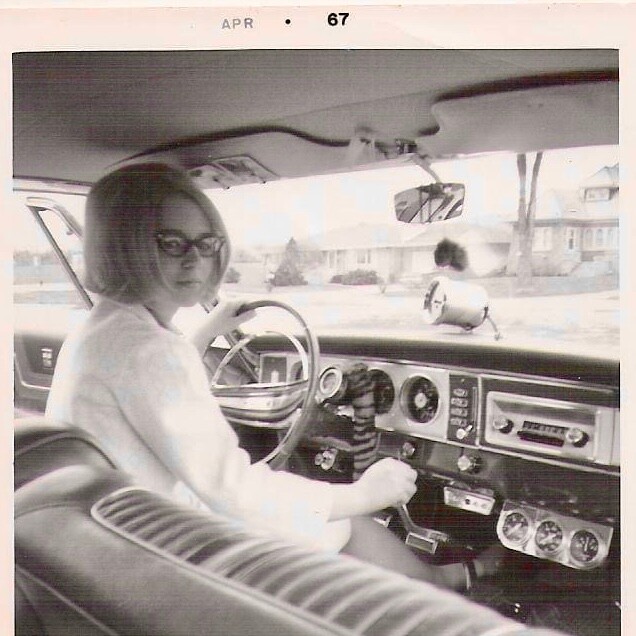 Woman with glasses sits in the driver's seat of a vintage car, hands on the steering wheel, retro dashboard gauges visible.
