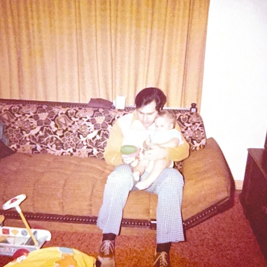Man sitting on a floral-patterned couch cradles a baby while holding a green toy. A cozy vintage living room scene appears behind them.