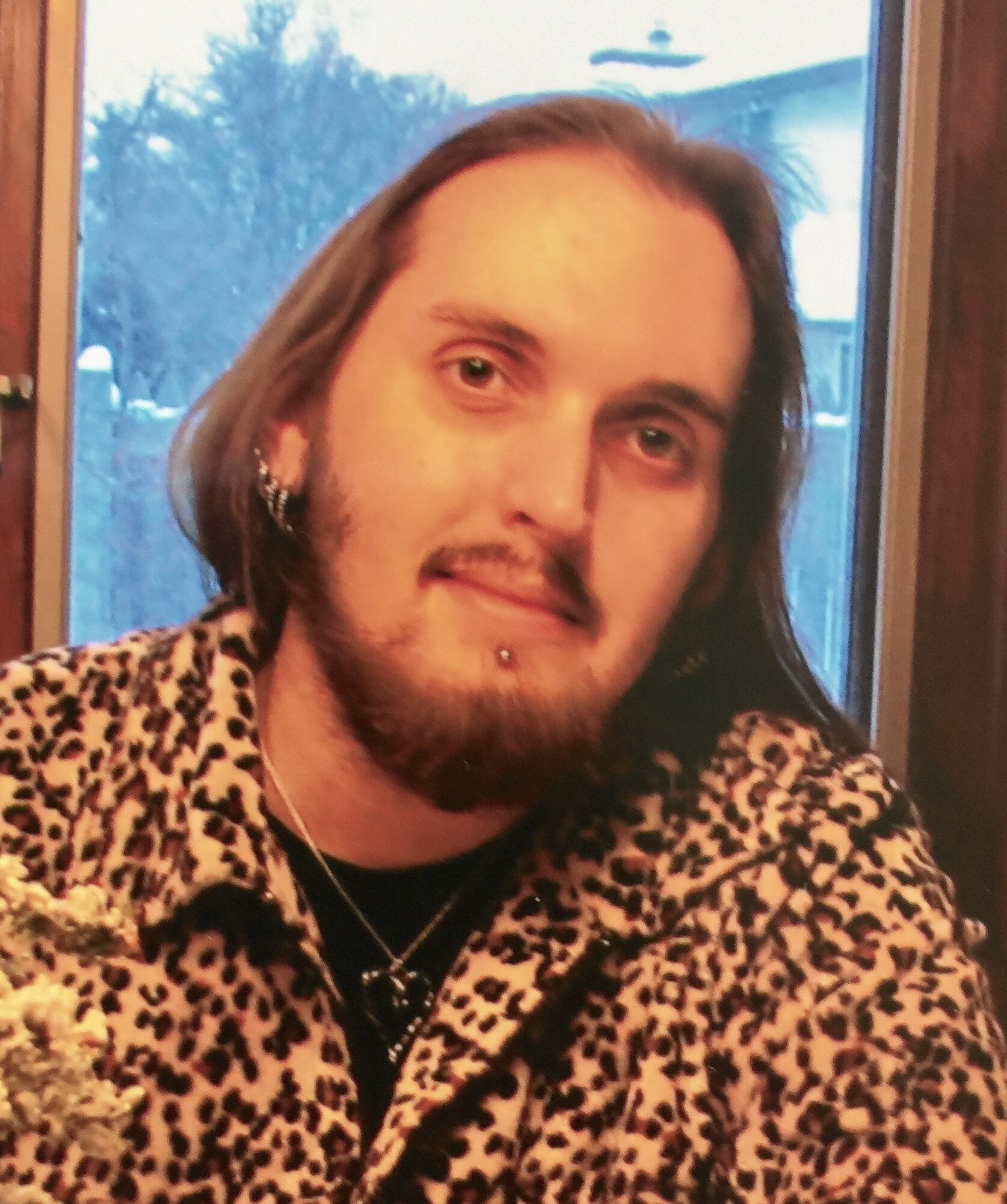 Close-up portrait of a man with shoulder-length hair and a beard wearing a leopard-print shirt inside a room.