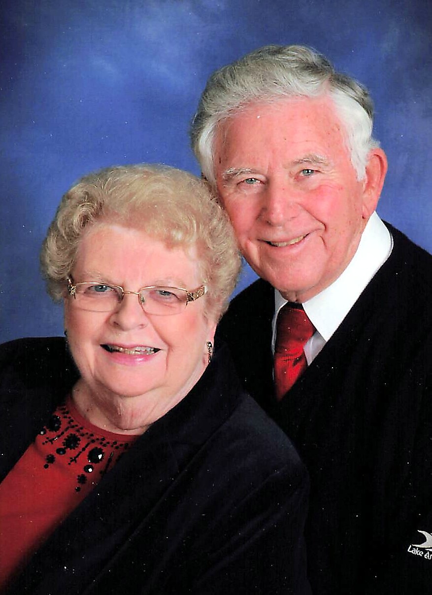 Smiling elderly couple posing for a studio portrait; woman wears glasses and a red blouse, man in a dark suit with a red tie against a blue backdrop.