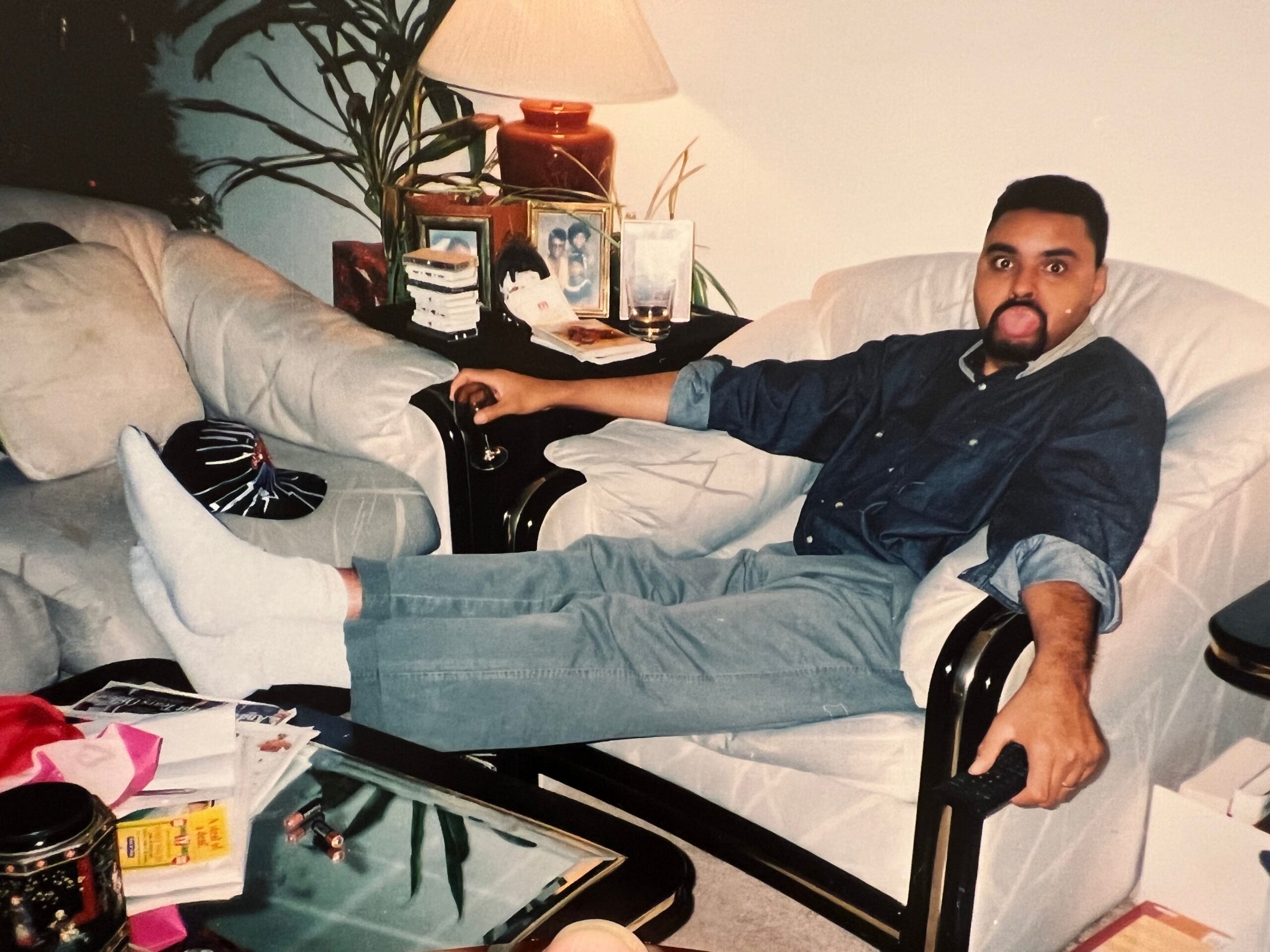Man on a sofa making a goofy face with tongue out, cluttered living room table behind him with photos, lamp, plant, and papers nearby.