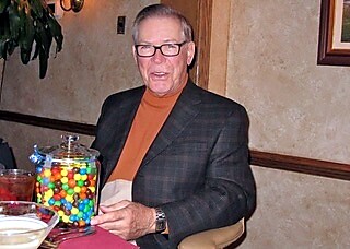Older man with glasses sits at a table indoors, wearing a dark blazer and orange turtleneck, with a jar of colorful candies in front of him.