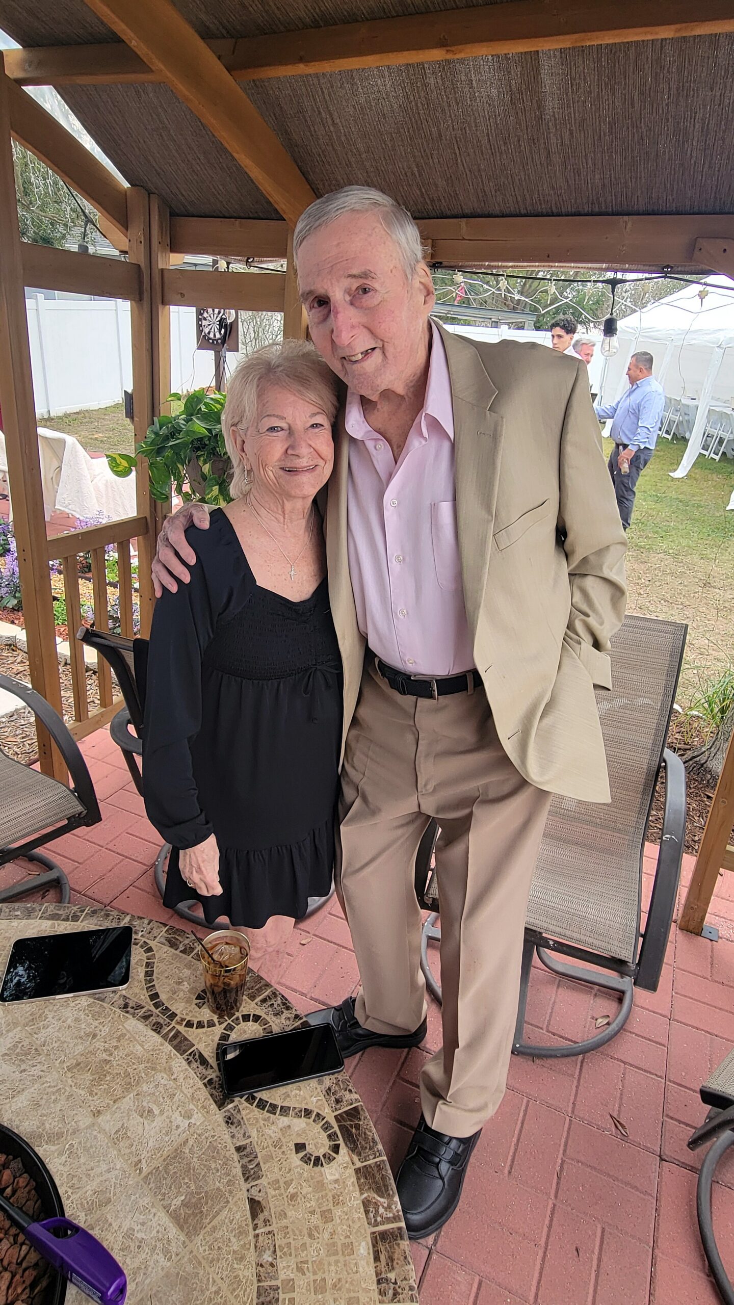 An elderly couple stands closely together outdoors under a wooden pergola, smiling at the camera; the man wears a tan suit with a pink shirt, wife in a black dress.