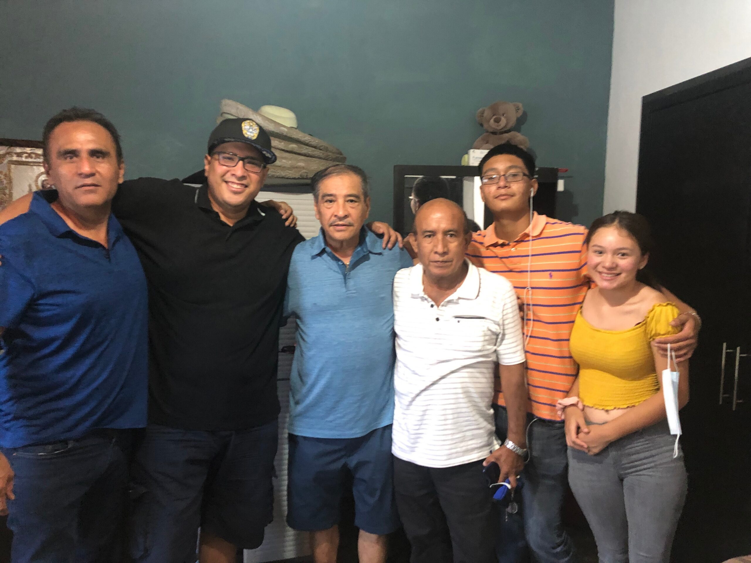 Group of seven people posing together indoors, smiling for the camera in casual clothes.