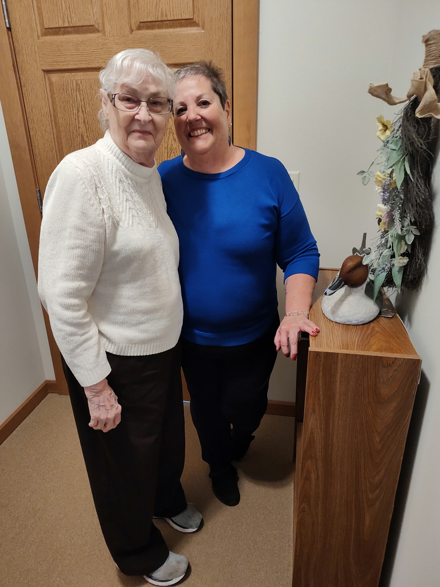 Two women posing together indoors: an older woman in a cream sweater and glasses beside a smiling woman in a royal blue top by a wooden cabinet and decorative arrangement.