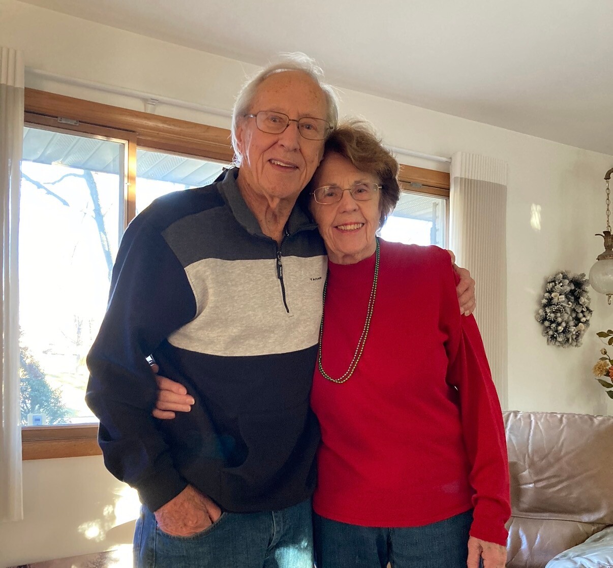 Two smiling elderly adults hug each other in a sunlit living room, wearing casual clothes (man in a dark sweater, woman in red).