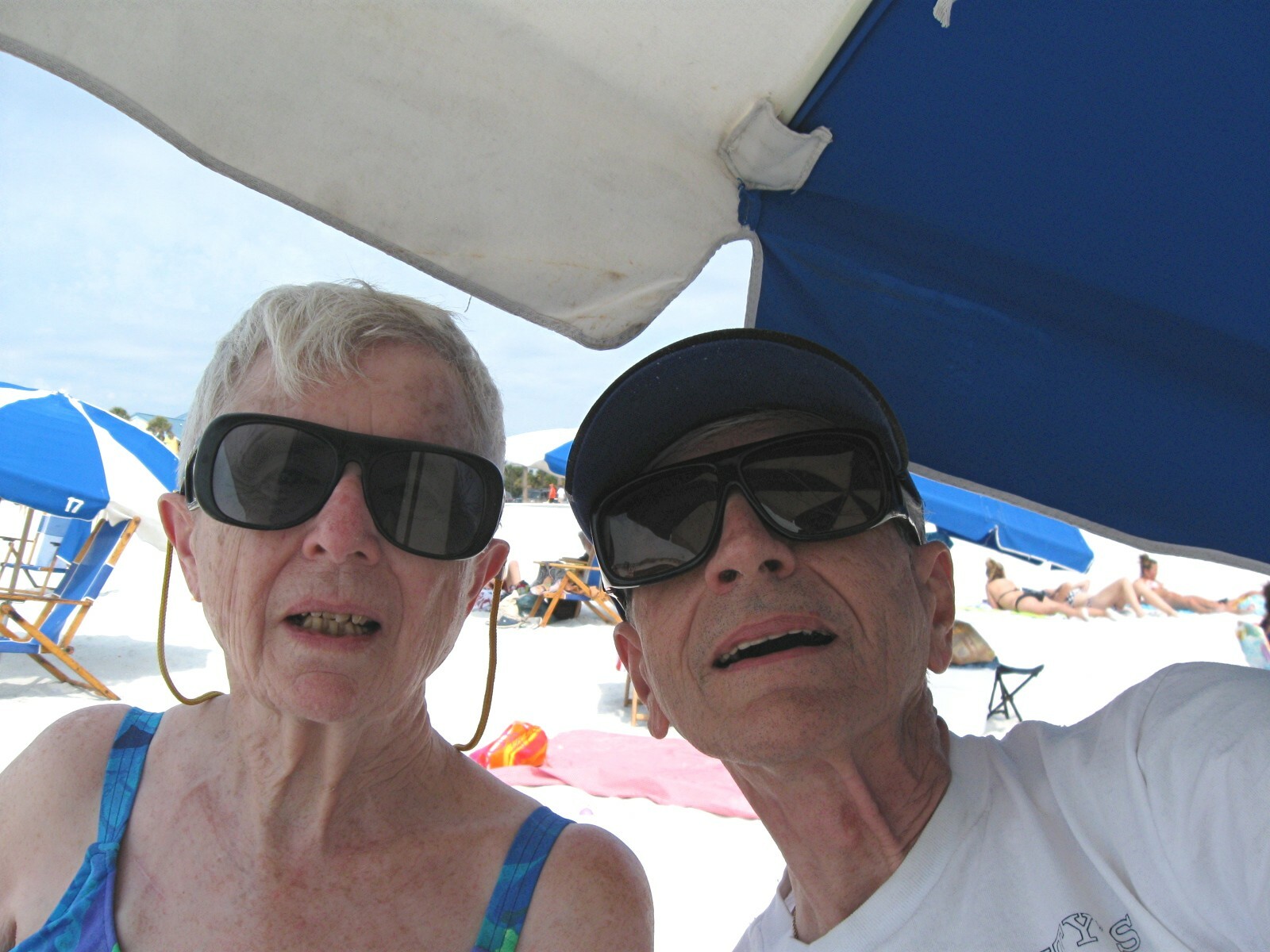 Two smiling older adults wearing sunglasses taking a selfie at the beach under a large umbrella.