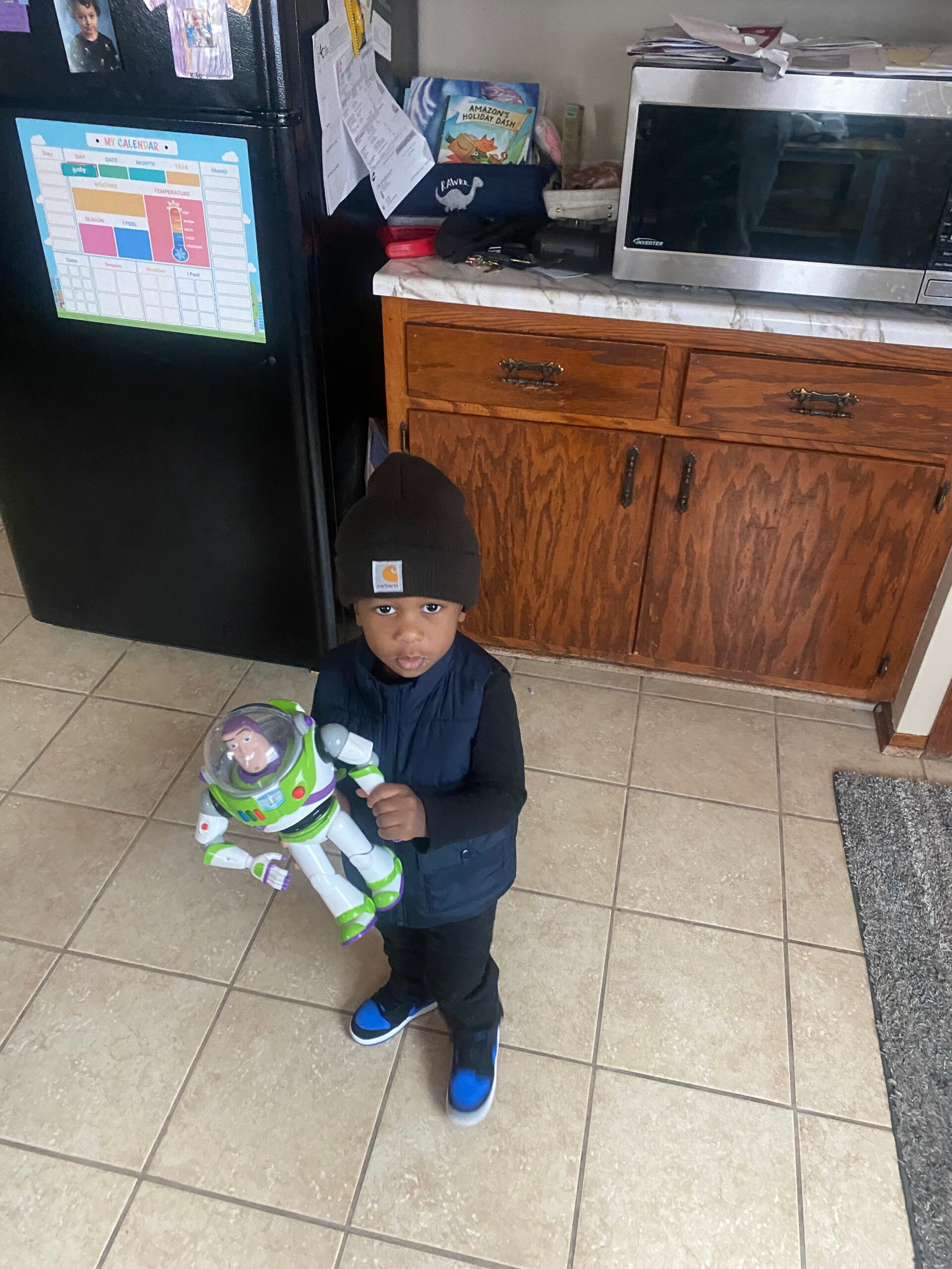 Young child in a dark beanie and navy vest holding a Buzz Lightyear toy in a kitchen by the refrigerator and microwave.