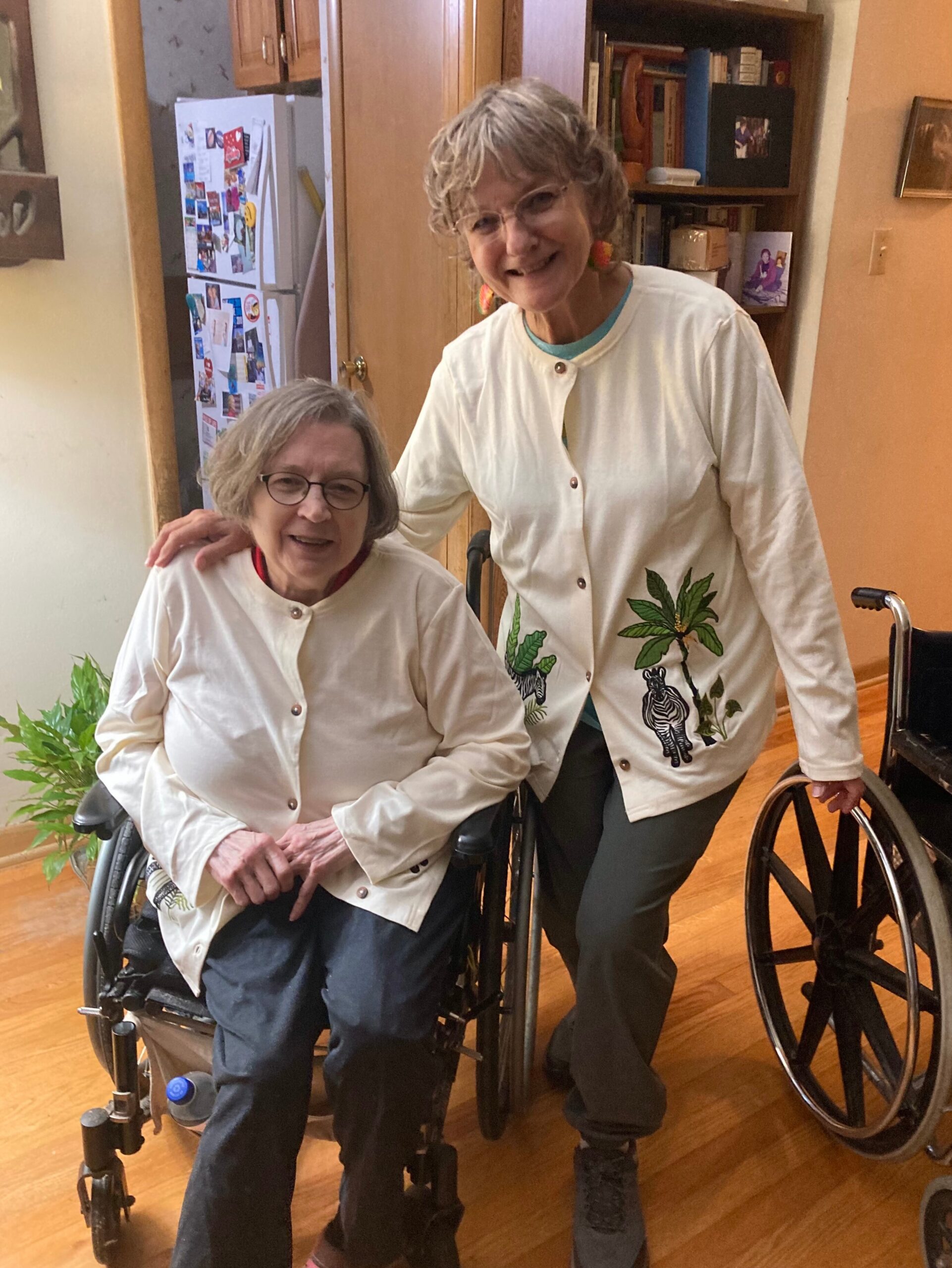 Two elderly women in wheelchairs indoors, one standing with arm around the other, both smiling in white jackets with tropical patterns.