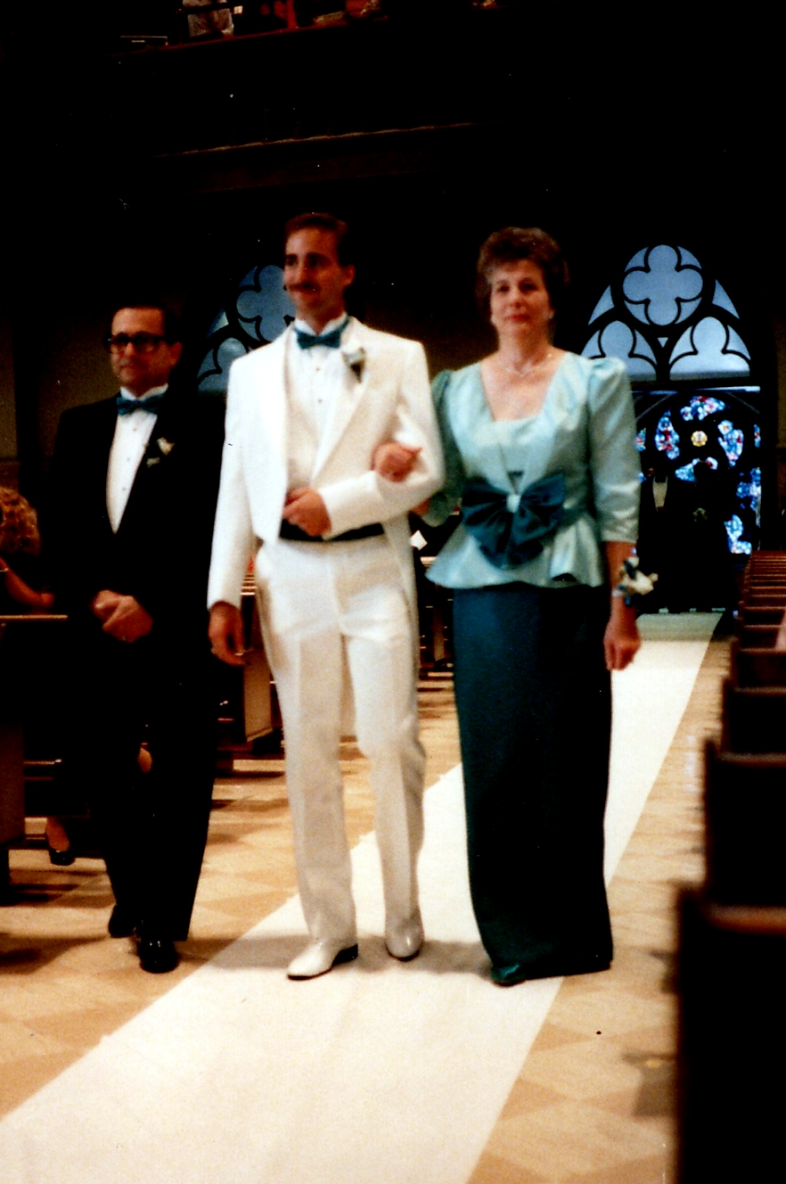 Three people in formal attire walk down a church aisle, likely part of a wedding procession. A man in a white tuxedo is escorted by a man in a dark suit on one side and a woman in a teal outfit on the other.