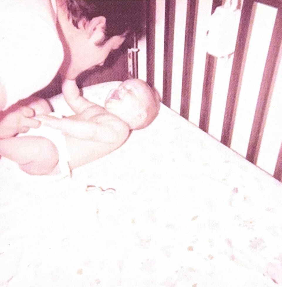 Newborn baby lying in a crib, one arm raised toward an adult hand reaching in from the left.