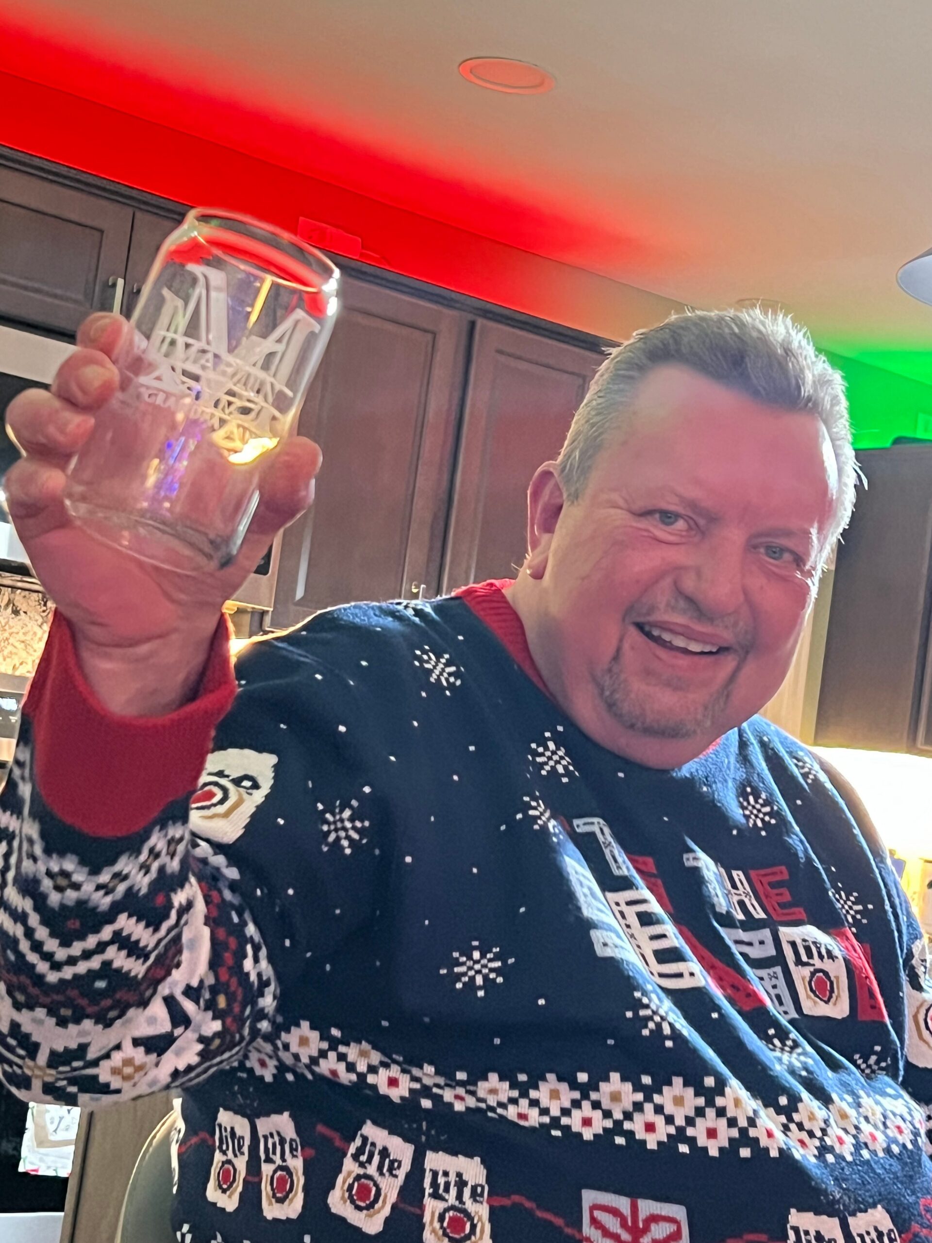 Smiling man in a blue holiday sweater raises a glass in a kitchen with red and green lighting.