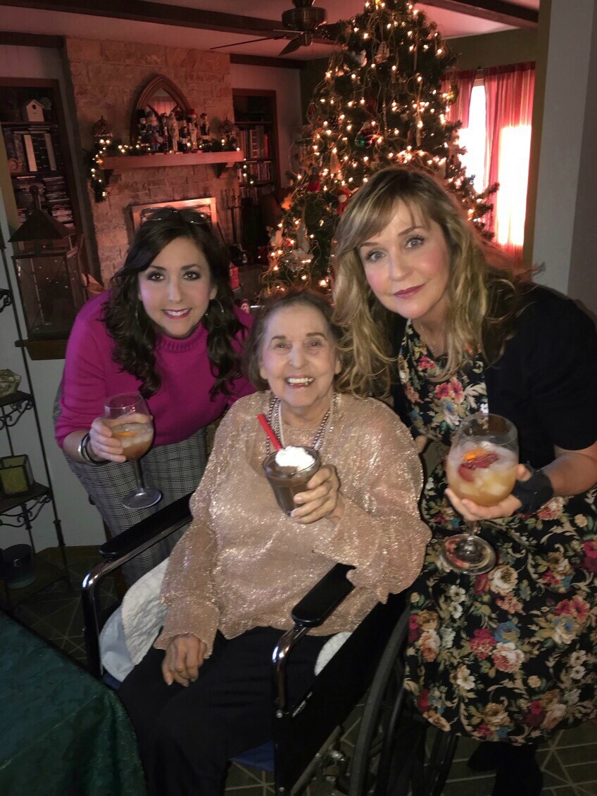 Three women smile and hold cocktails around a Christmas tree; elderly woman in a wheelchair at center wearing a sparkly pink top.