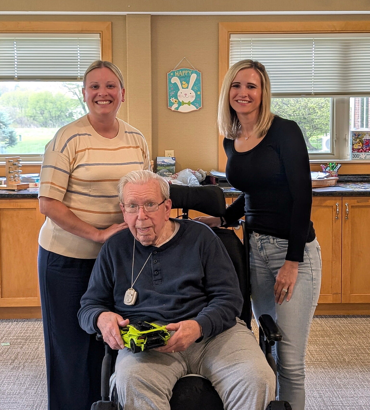 Three smiling adults with one elderly man in a wheelchair holding a green toy car, indoors near cabinets and windows with a bunny sign in the background