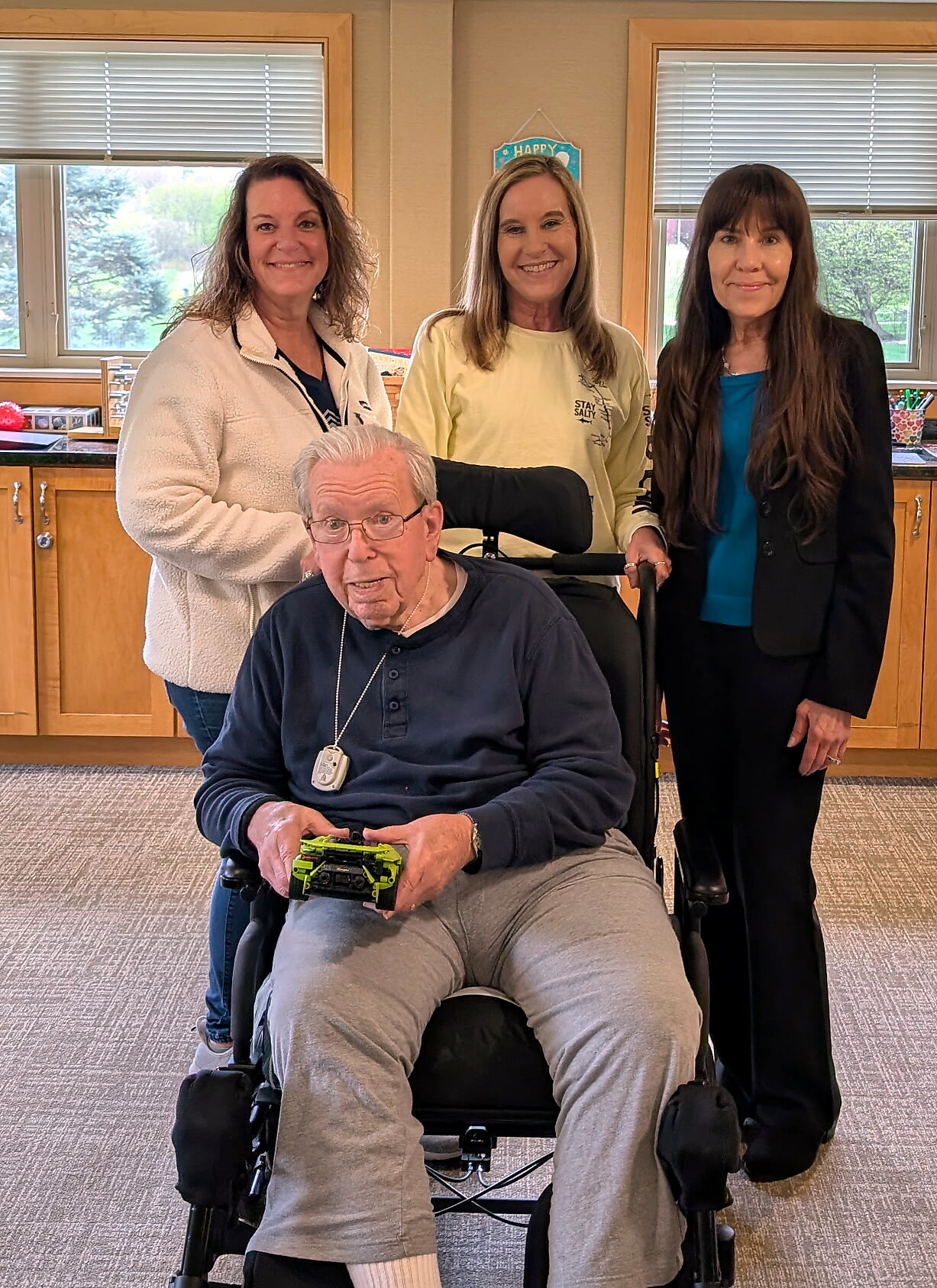 Older man in a motorized wheelchair in front, four adults standing behind him smiling in a room with windows.