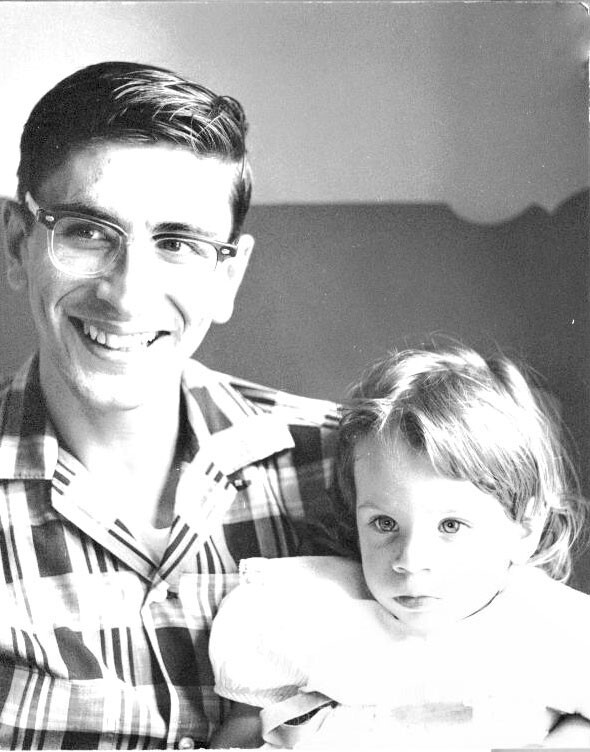 Smiling man in a plaid shirt with a young child on his lap in a black-and-white photo.