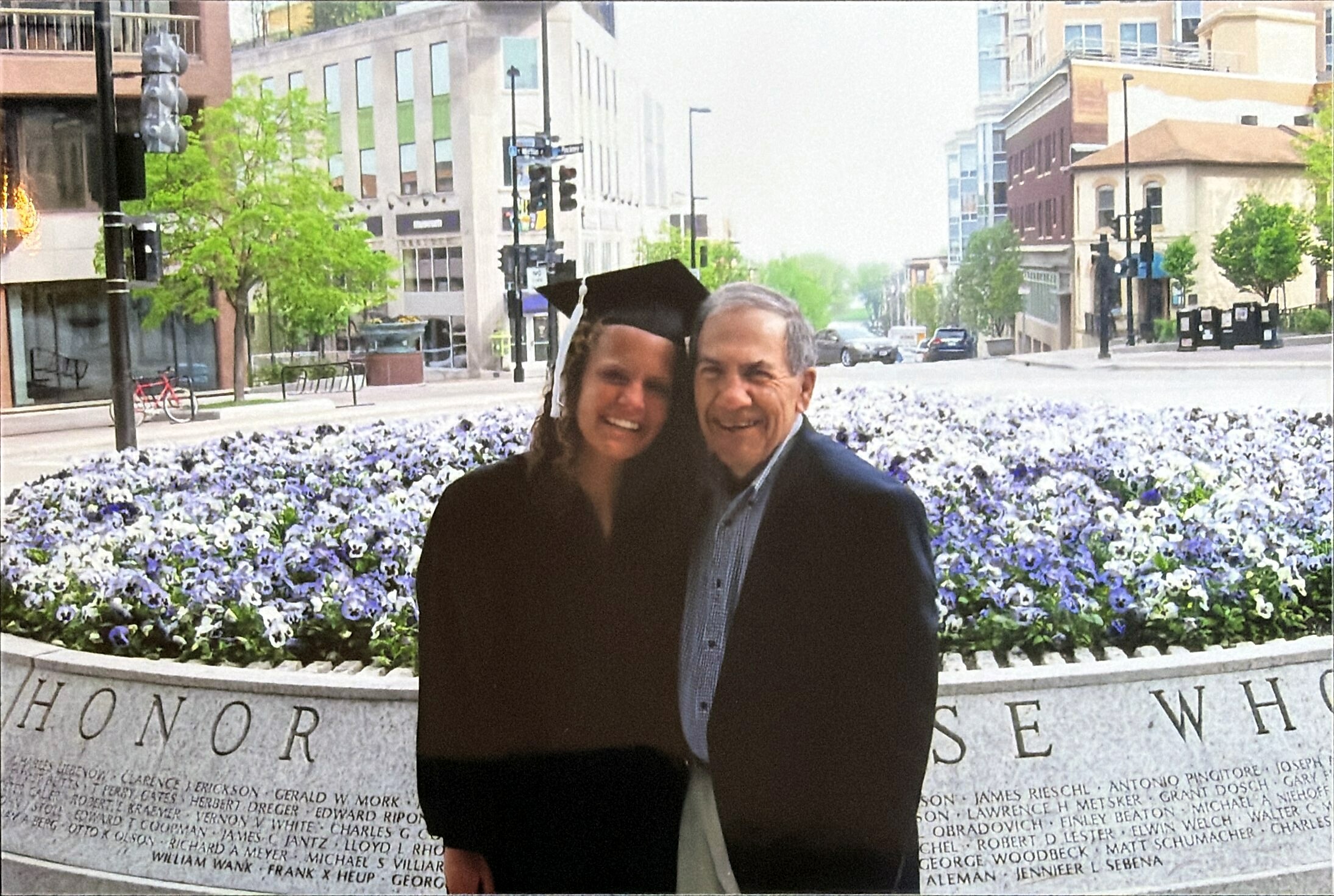 Smiling graduate in a cap and gown posing with an older man in front of a name-inscribed memorial wall and purple flower bed on a city street.