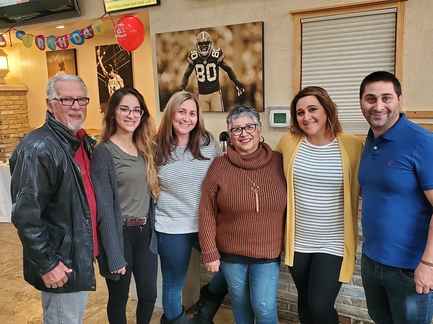 Six adults posing together indoors at a birthday party, smiling, with a colorful banner and red balloon in the background.