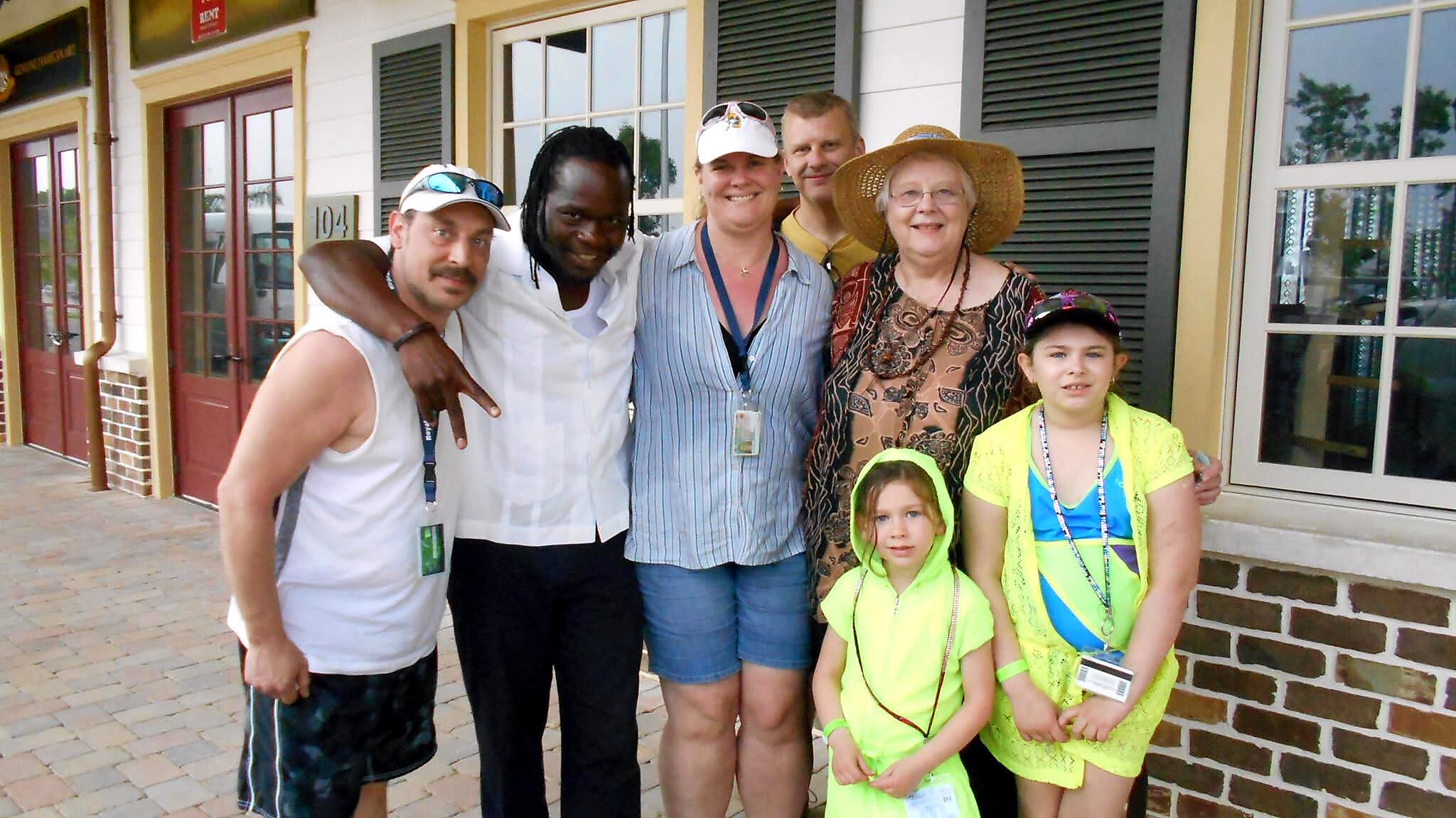 A diverse group of six people posing together outside a brick storefront, smiling at the camera, two kids in neon yellow outfits at the front.