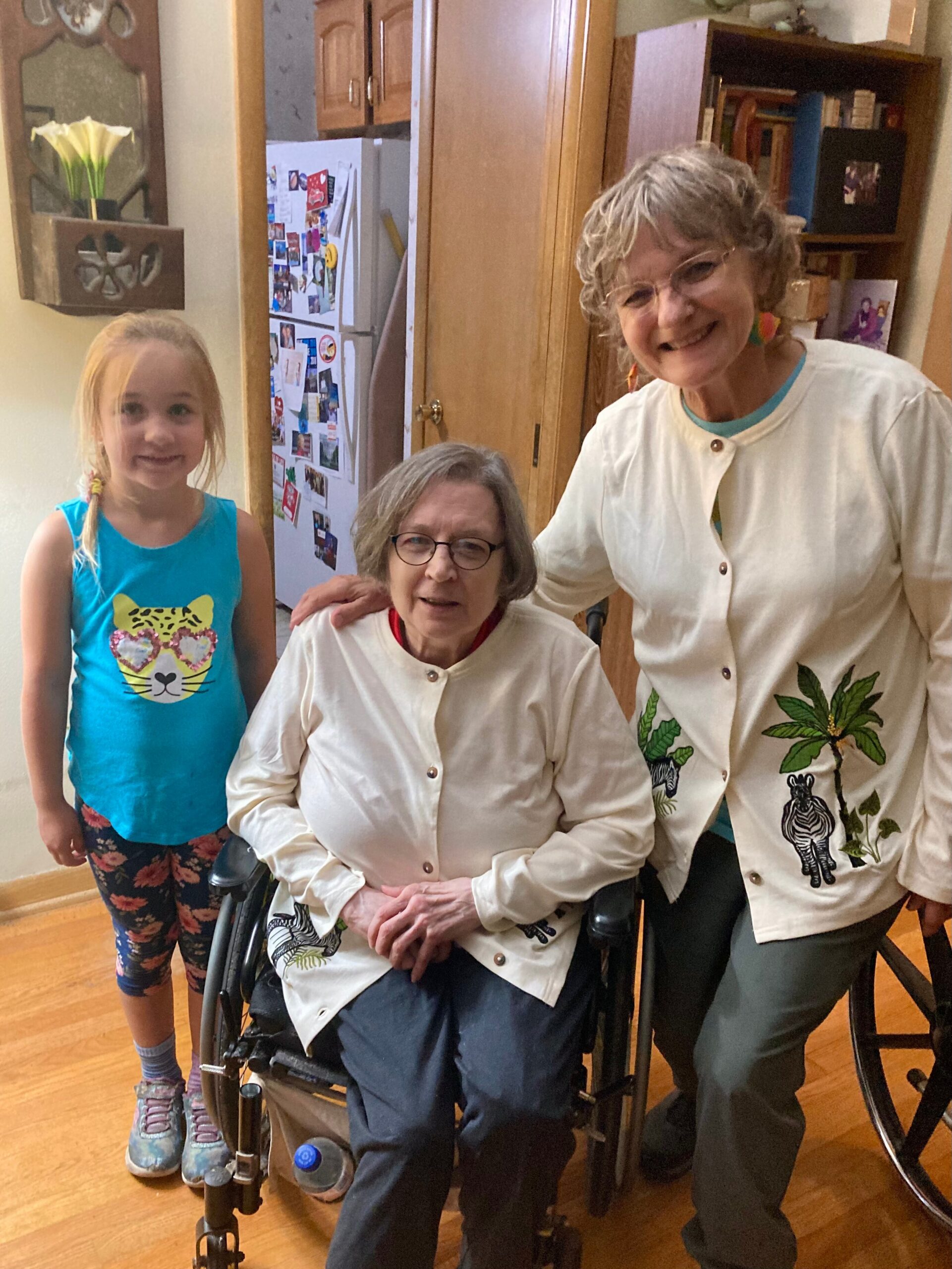 A young girl in a blue sleeveless shirt stands beside two smiling older women in a kitchen; one is in a wheelchair.