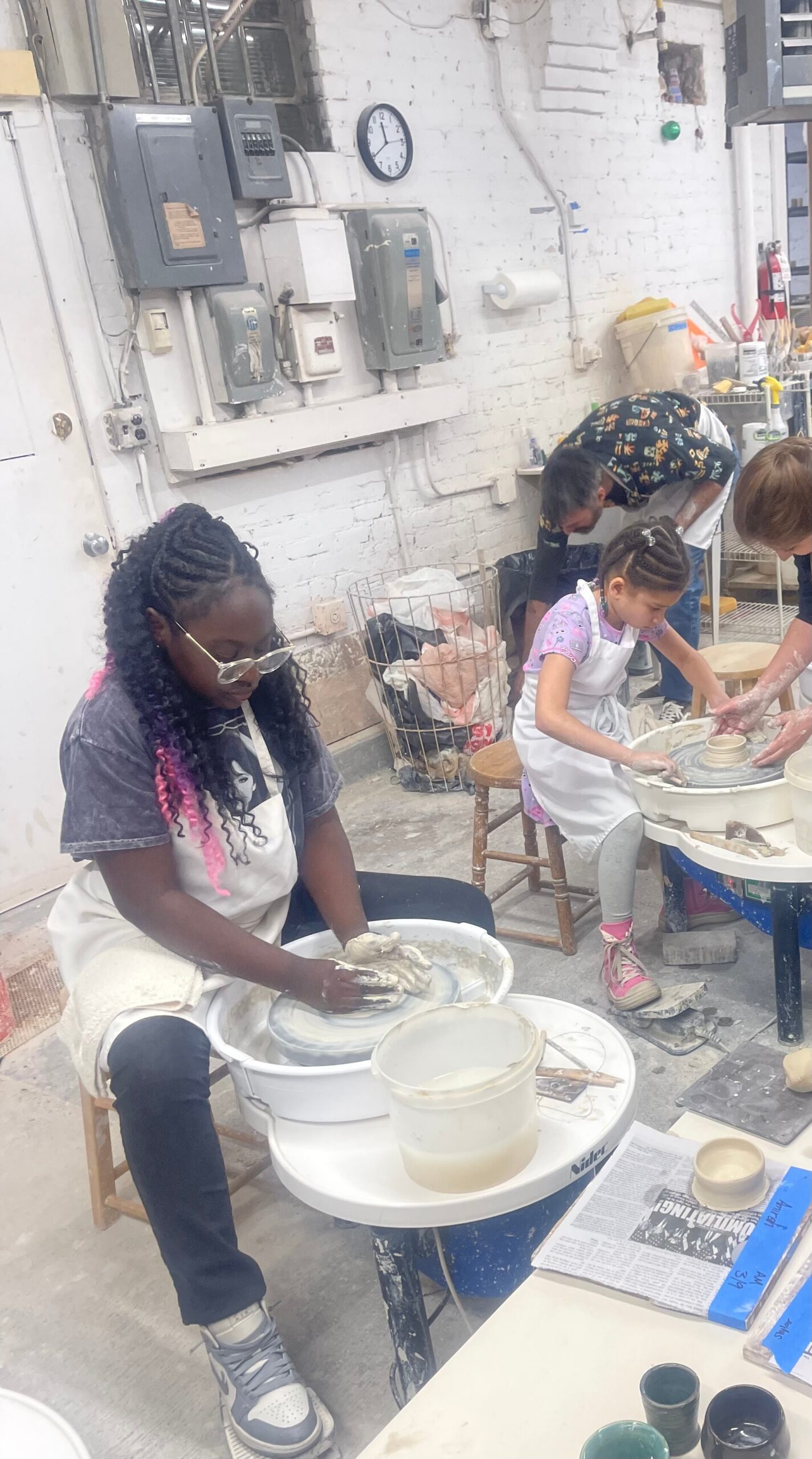 Children at pottery wheels shaping clay in a bright workshop with white brick walls and supplies nearby.