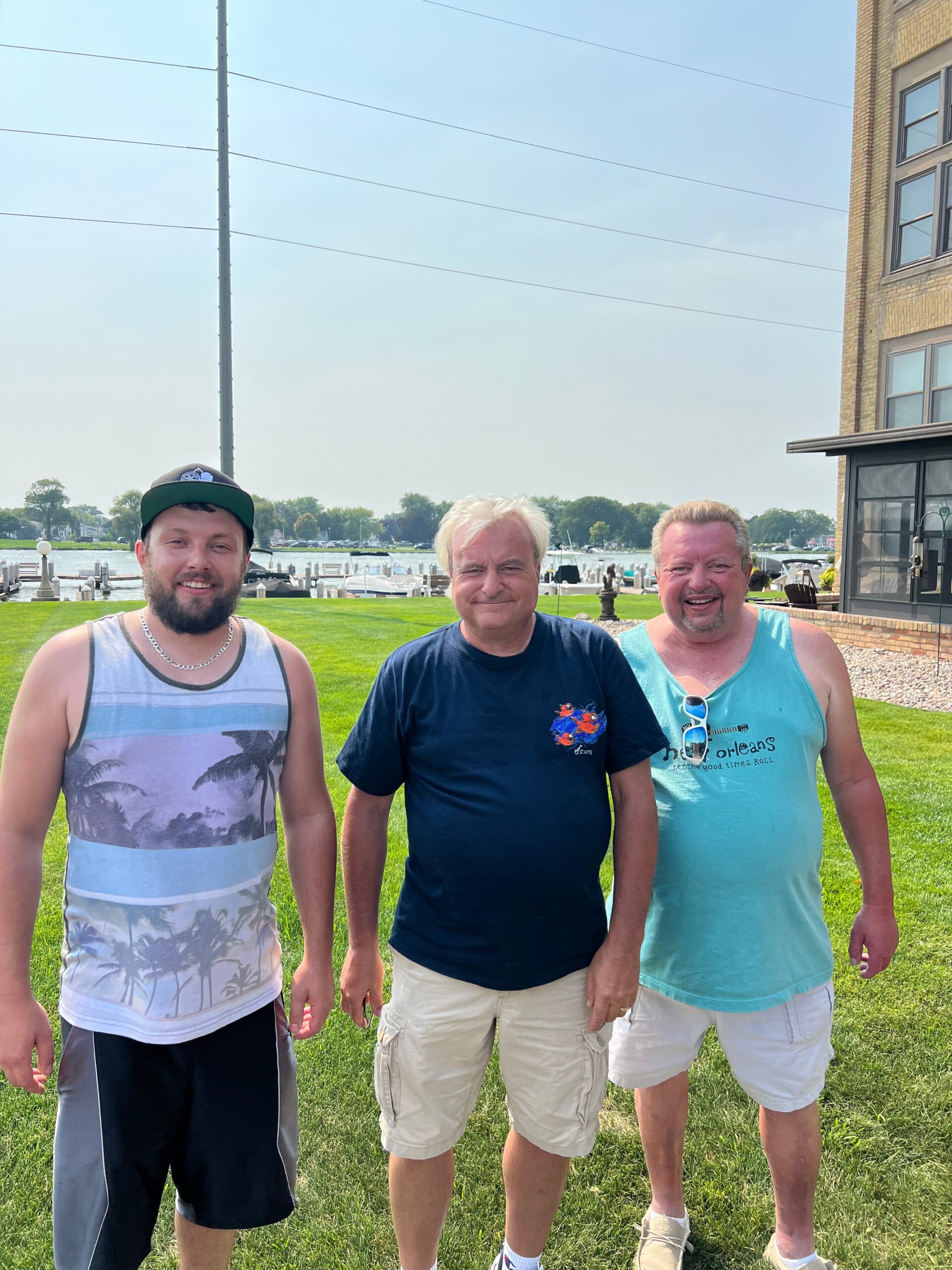 Three men stand side by side outdoors on a grassy area near a marina, posing for a photo with water and boats in the background.