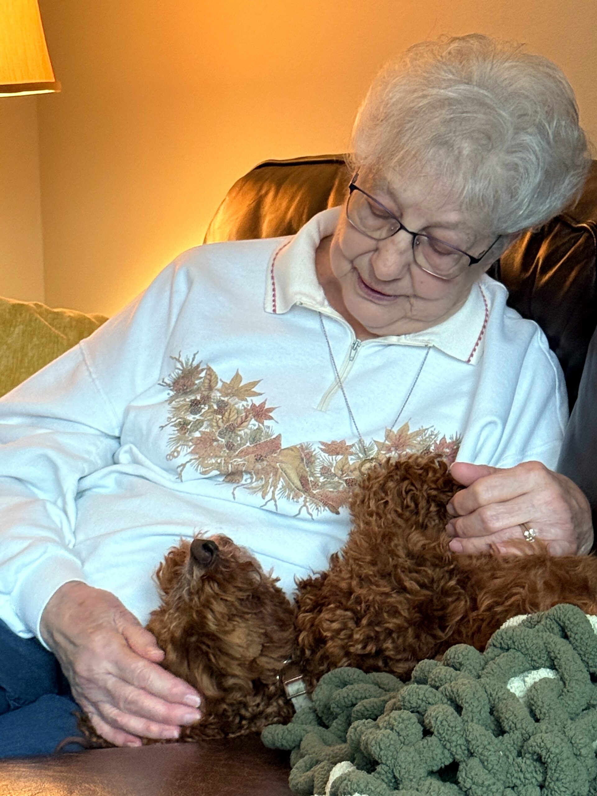 Senior woman with short gray hair and glasses sits on a leather sofa, smiling as she pets a fluffy brown dog, with a warm lamp behind her and a green crocheted blanket in the foreground.