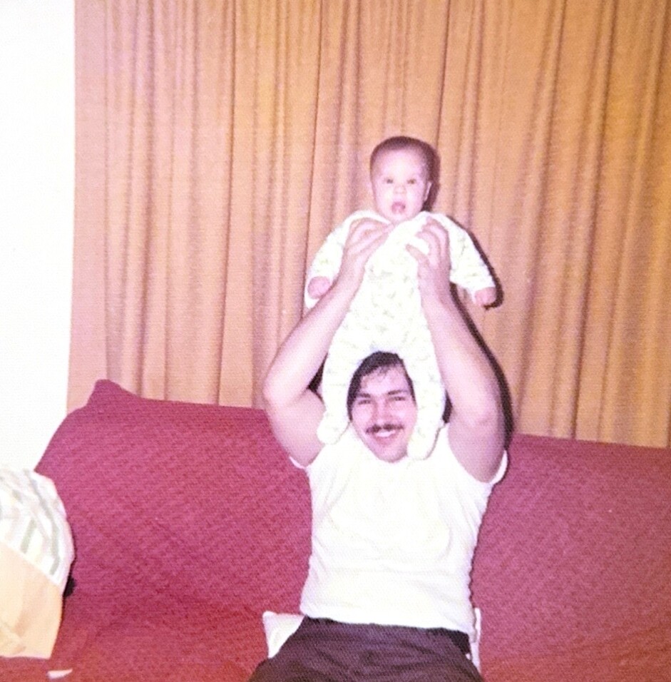 A man with a mustache lifting a baby above his head while sitting on a red couch indoors, yellow curtains in the background.