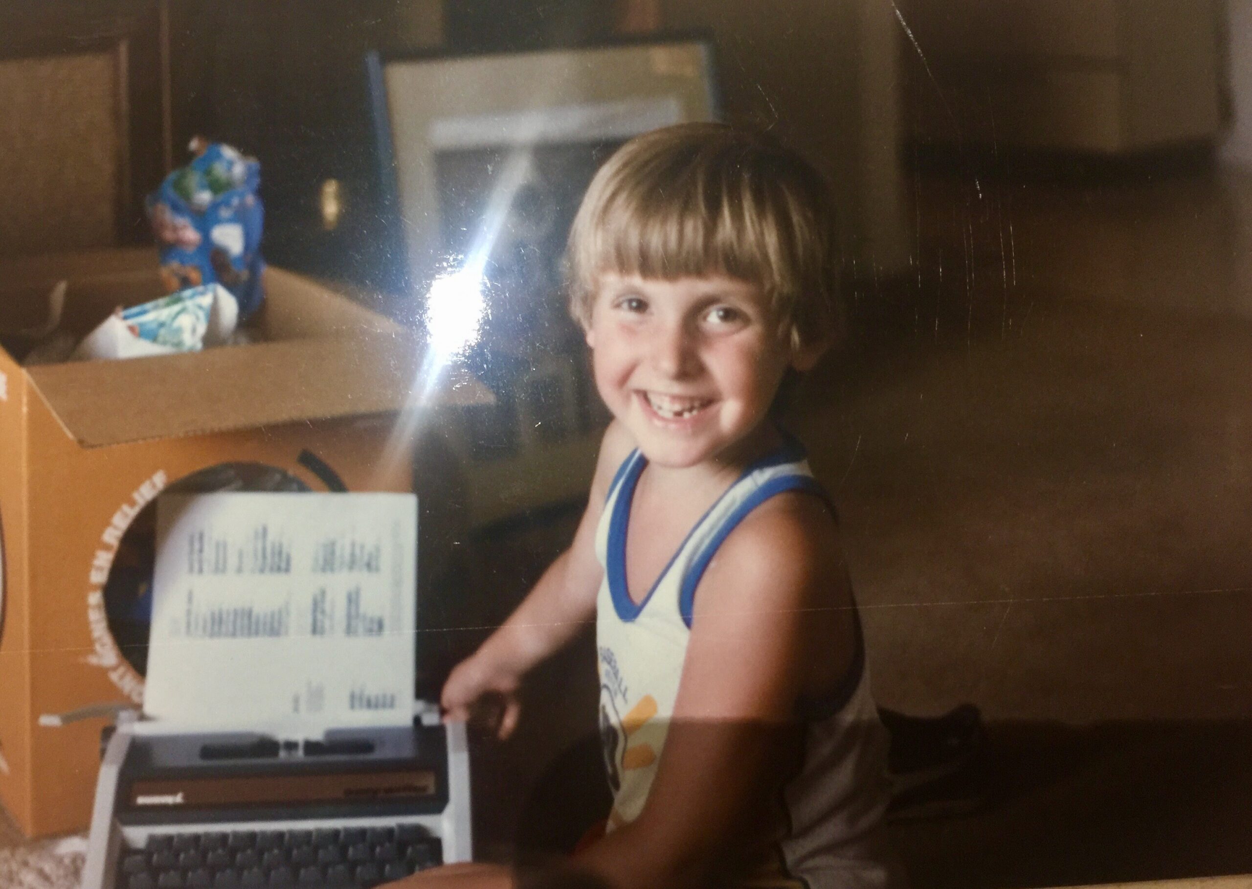 Smiling boy with blond hair in a blue-and-white striped shirt sits beside a vintage desktop computer/printer.