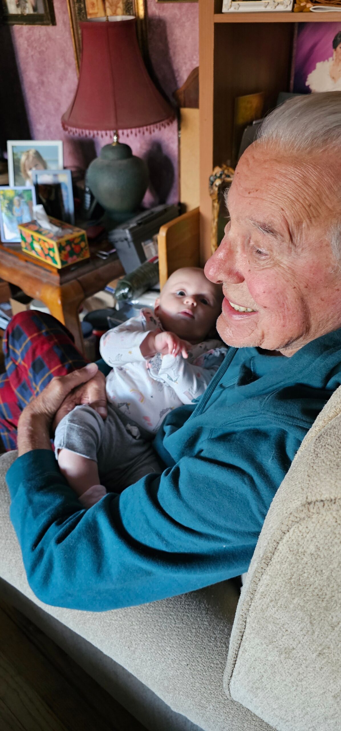 Grandfather cuddling a baby on a beige sofa in a cozy living room, both smiling softly at the camera.