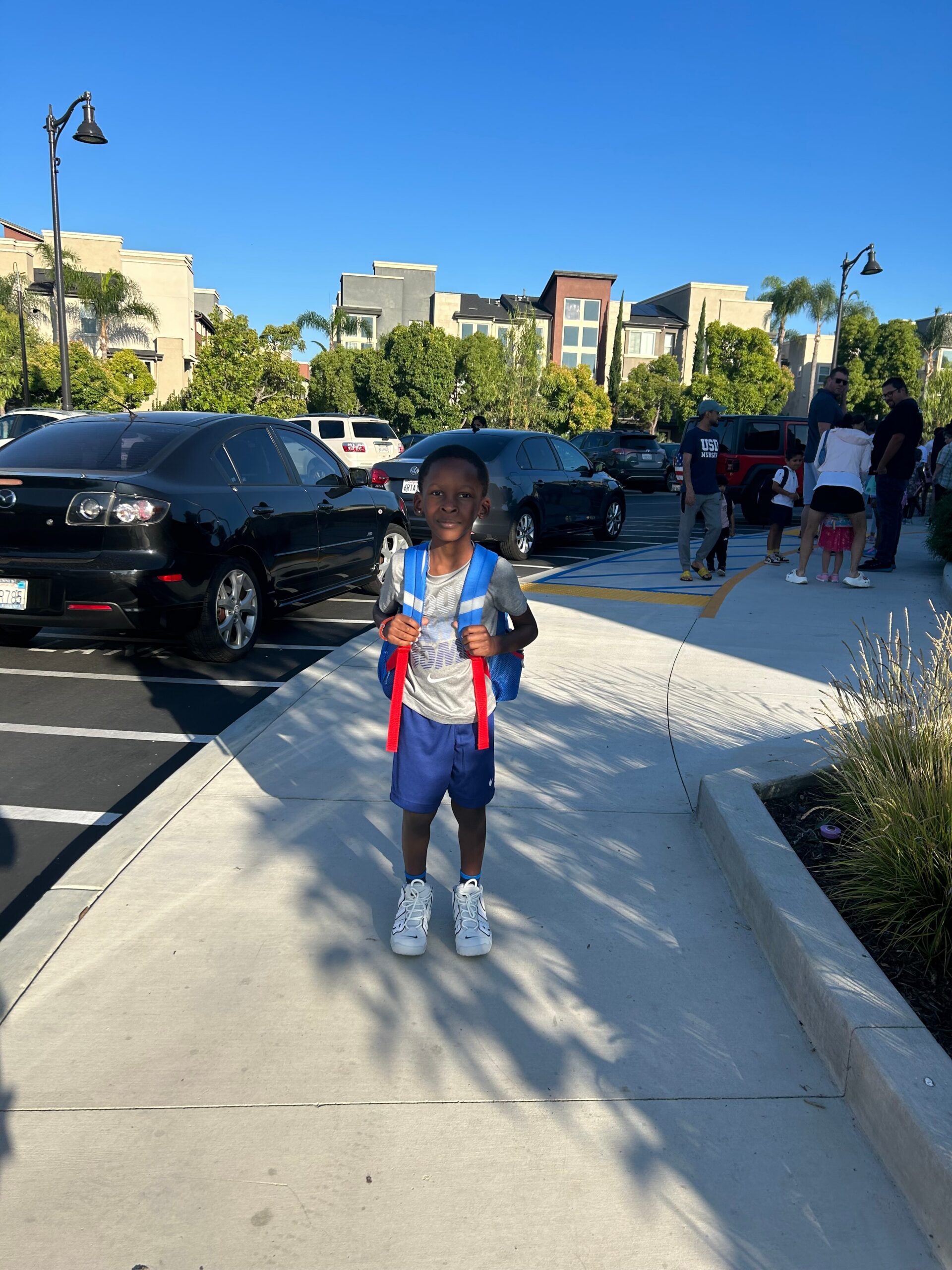 Young boy with a blue backpack standing in a parking lot on a sunny day, with cars and a group of people in the background.