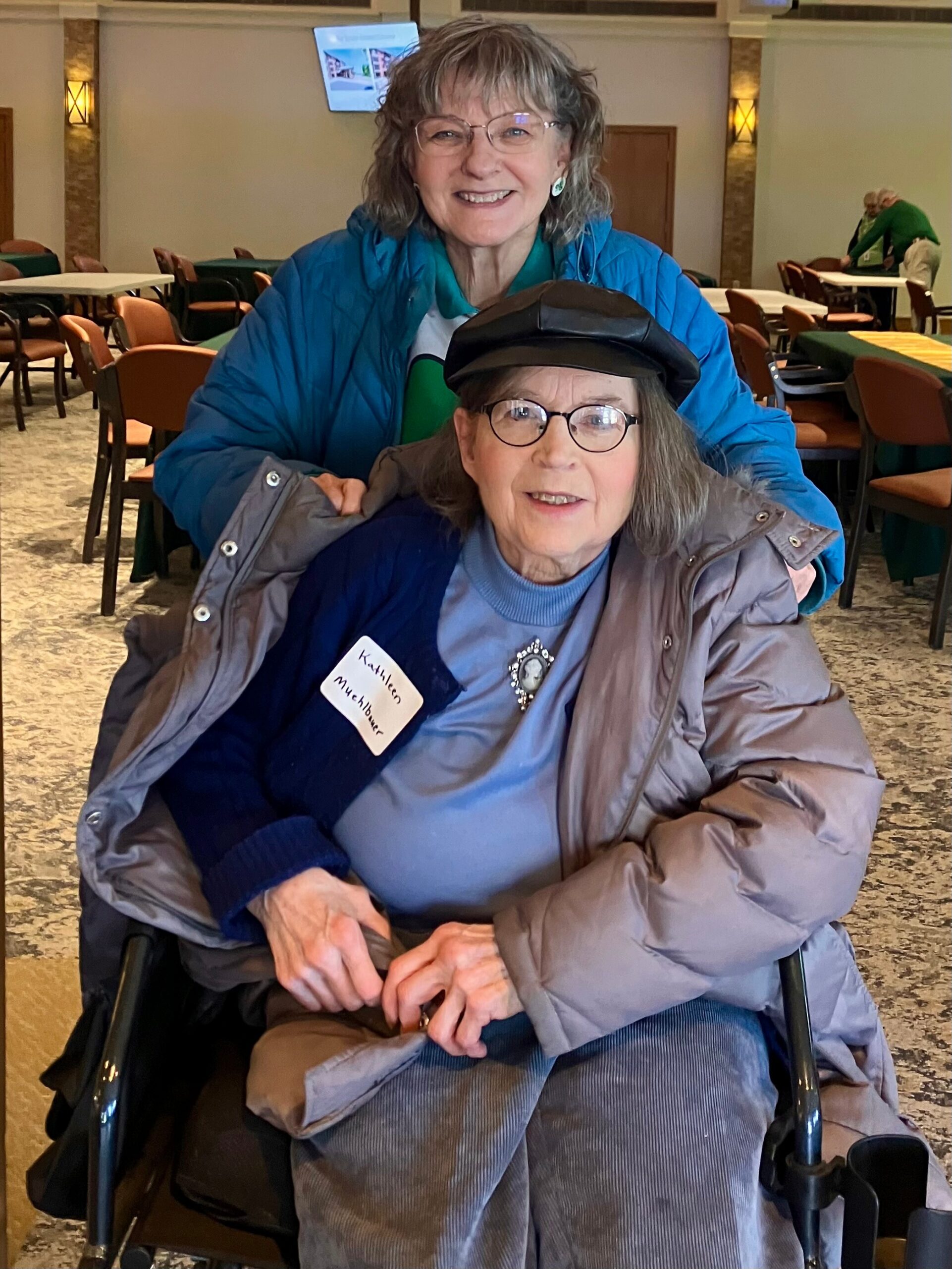 Two smiling women in a cafe, one seated in a wheelchair wearing a brown jacket and blue sweater, the other standing behind her in a bright blue jacket.