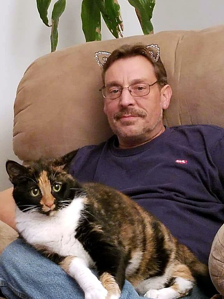 Man wearing glasses and a cat-ear headband sits on a beige recliner with a calico cat lounging on his lap, both facing the camera.