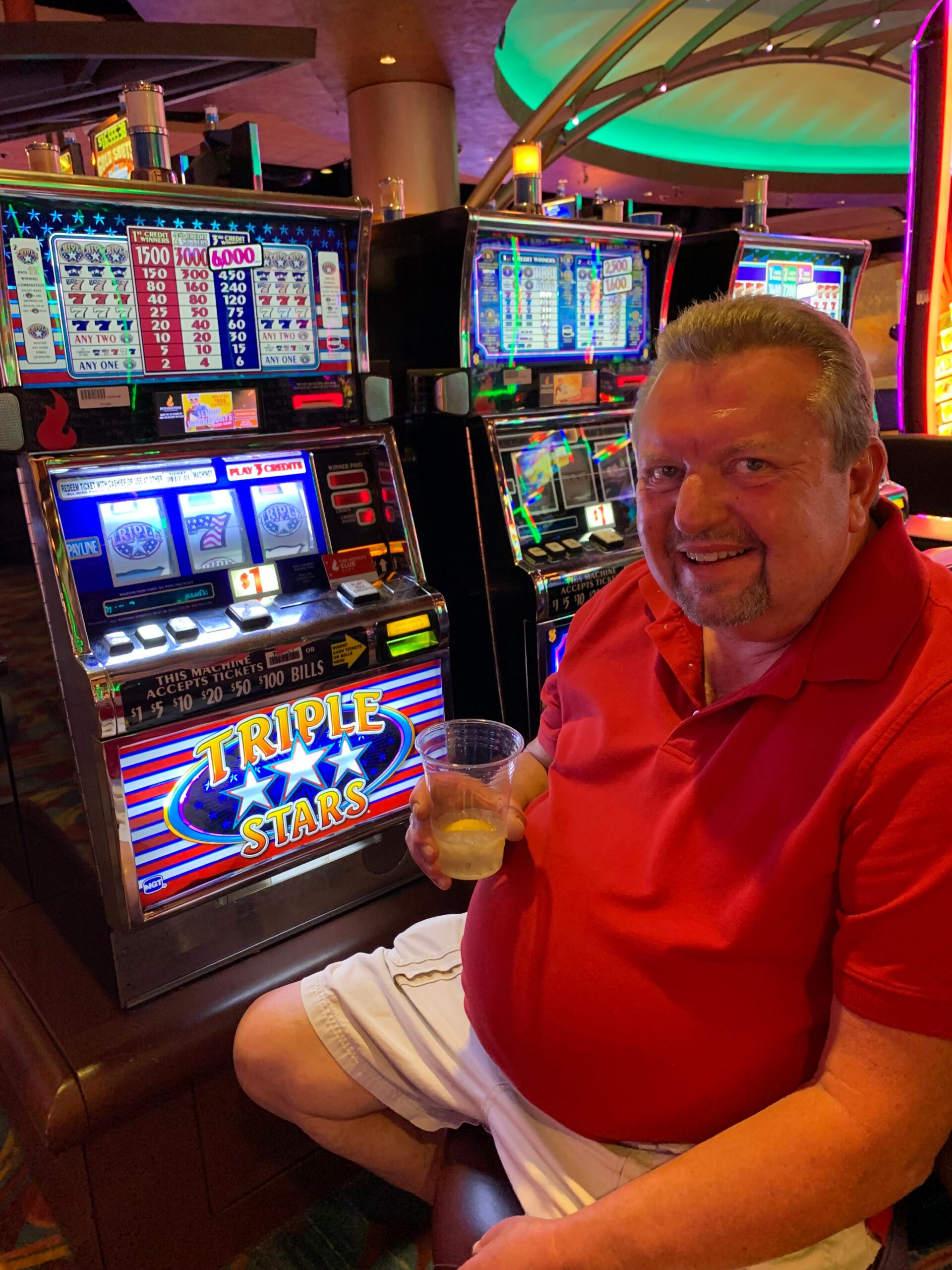 Smiling man in a red polo shirt sits at a row of bright slot machines, holding a plastic cup in a casino.