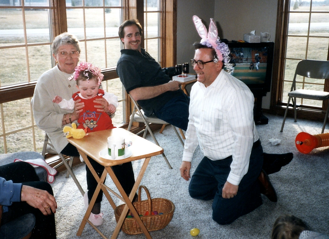 Family indoor gathering: an elderly woman holds a baby girl in a red outfit as others relax by large windows, wearing playful bunny ears for a festive moment.