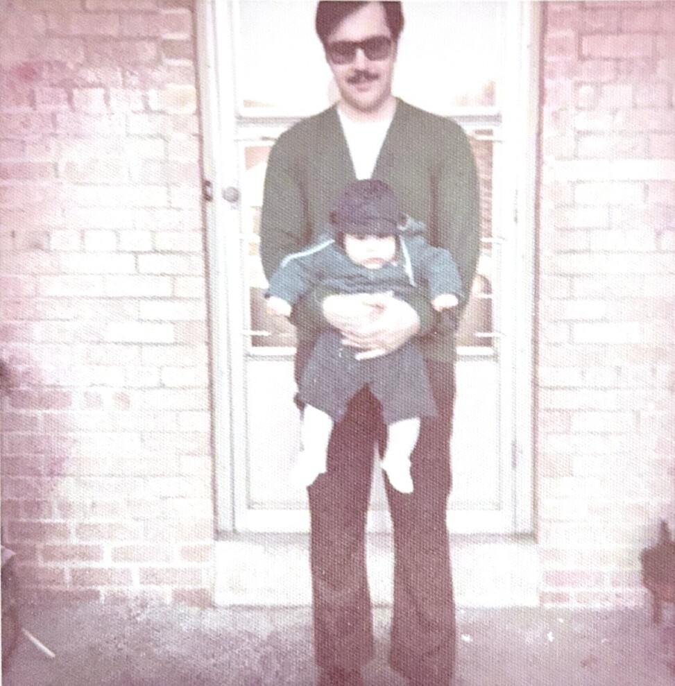 Man wearing sunglasses and a mustache, standing at a brick doorway, holding a small child dressed in dark clothes.