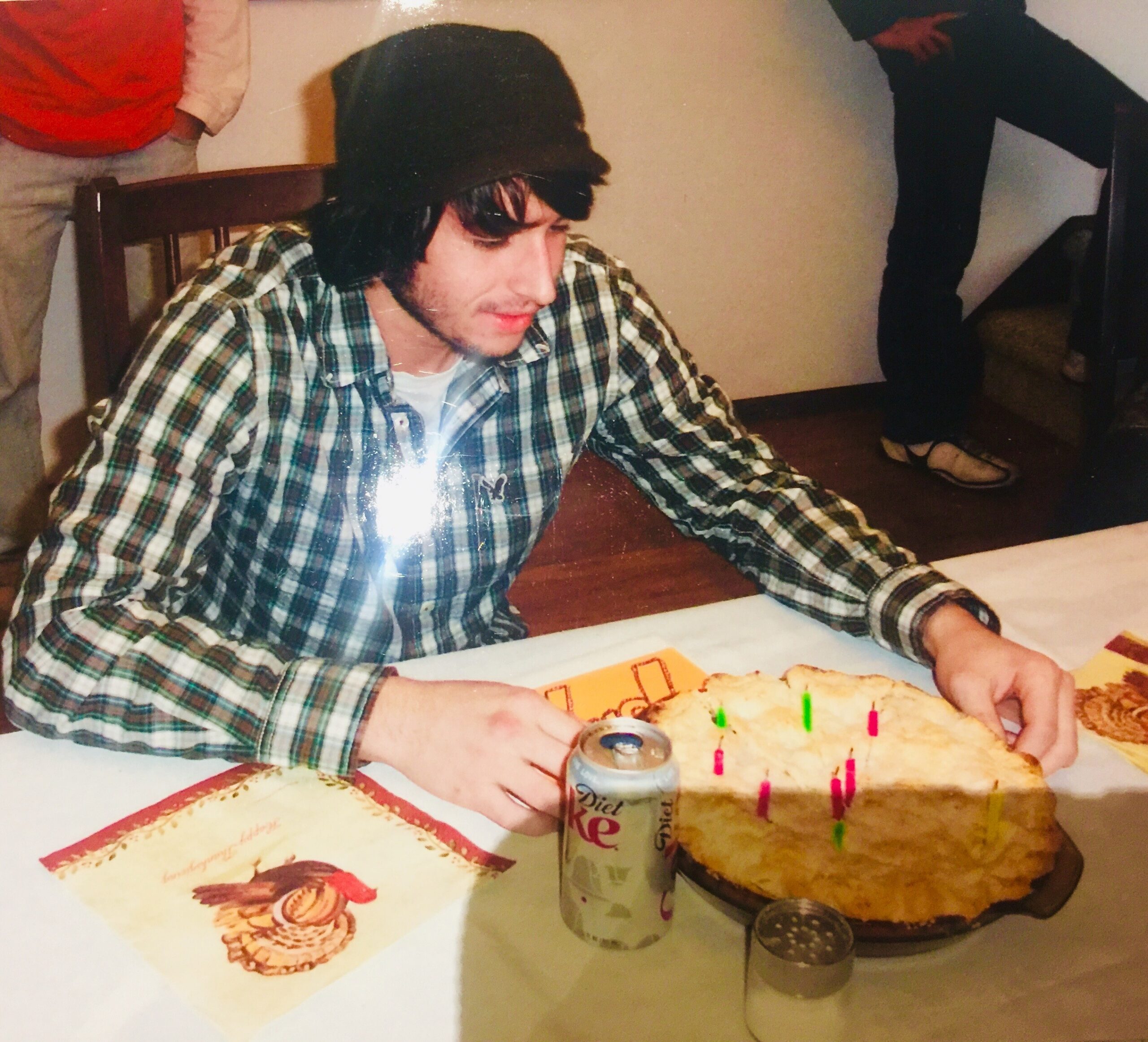 Young man with dark hair in a plaid shirt sits at a table with a cake and lit pink candles; a soda can nearby, others in the background.